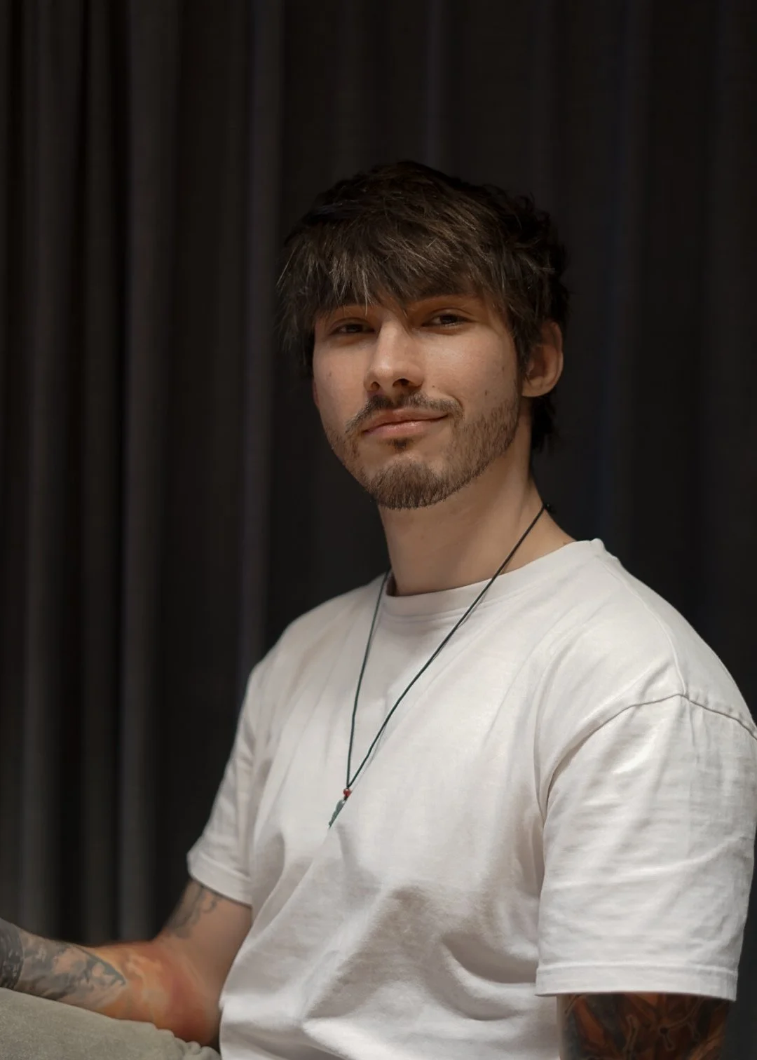A young man with wavy brown hair, a beard, and tattoos on his arms, wearing a white t-shirt and a necklace, sitting against a dark curtain background.