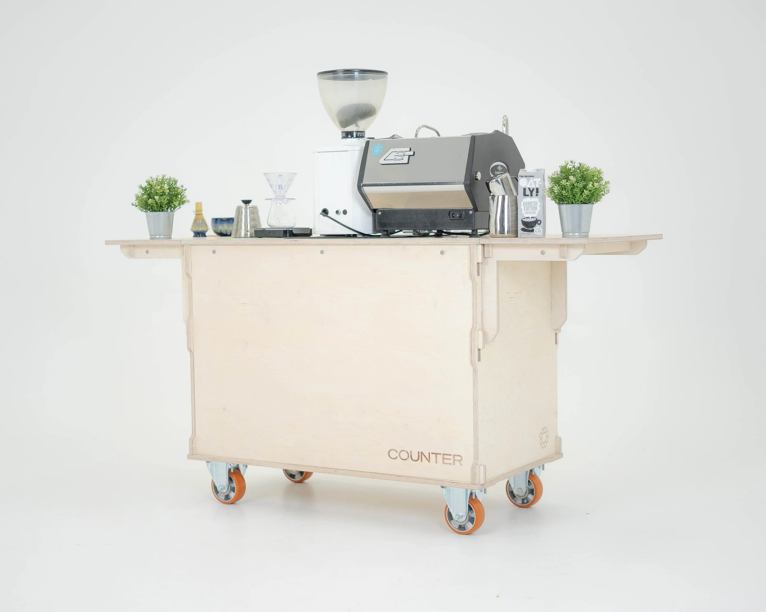 Coffee brewing station with coffee machine, grinder, and brewing accessories on a wooden mobile counter with plants on each side.