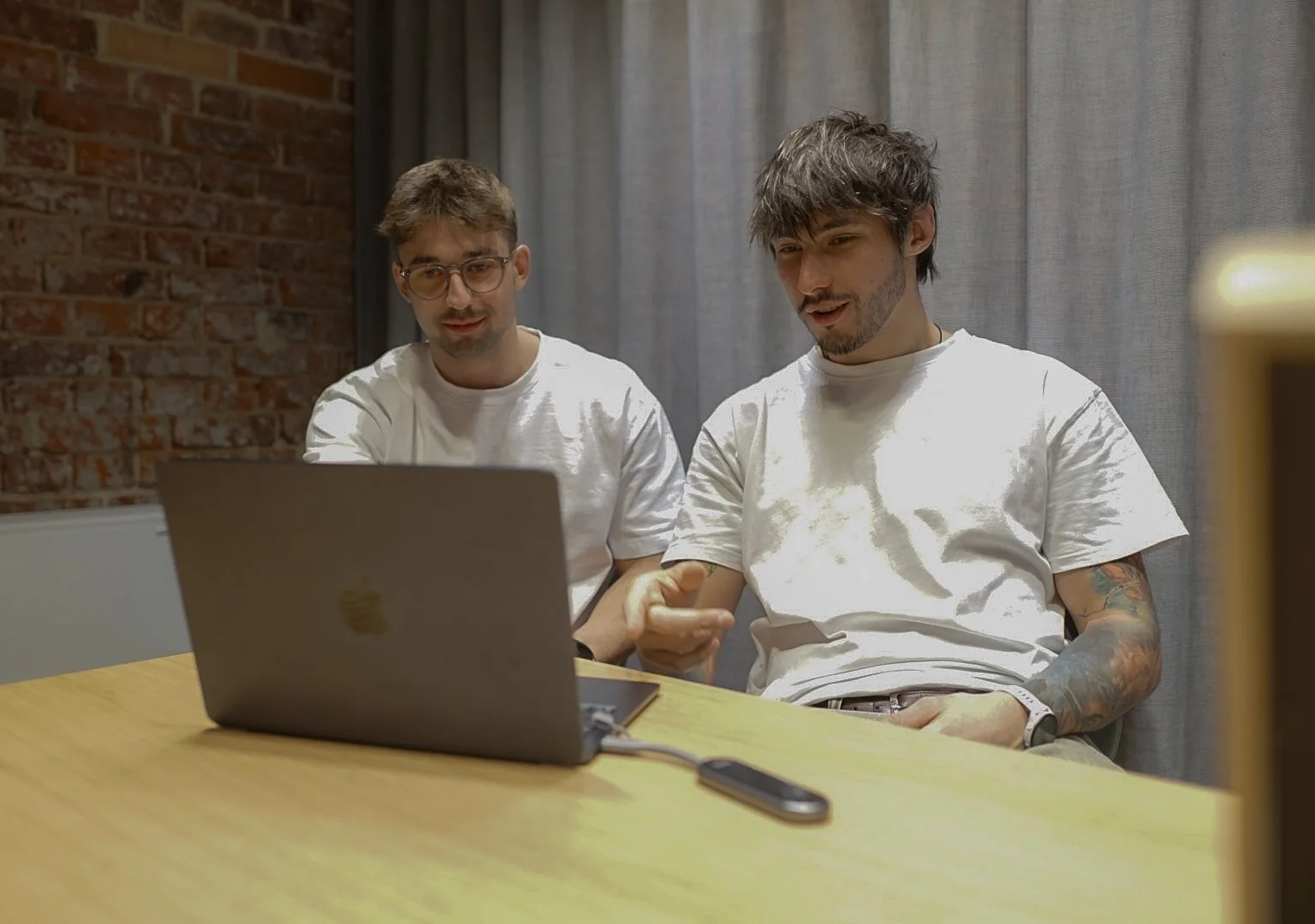 Two young men sit at a wooden table looking at a laptop screen, with one pointing at the screen. They are in an indoor space with a brick wall and gray curtains in the background.