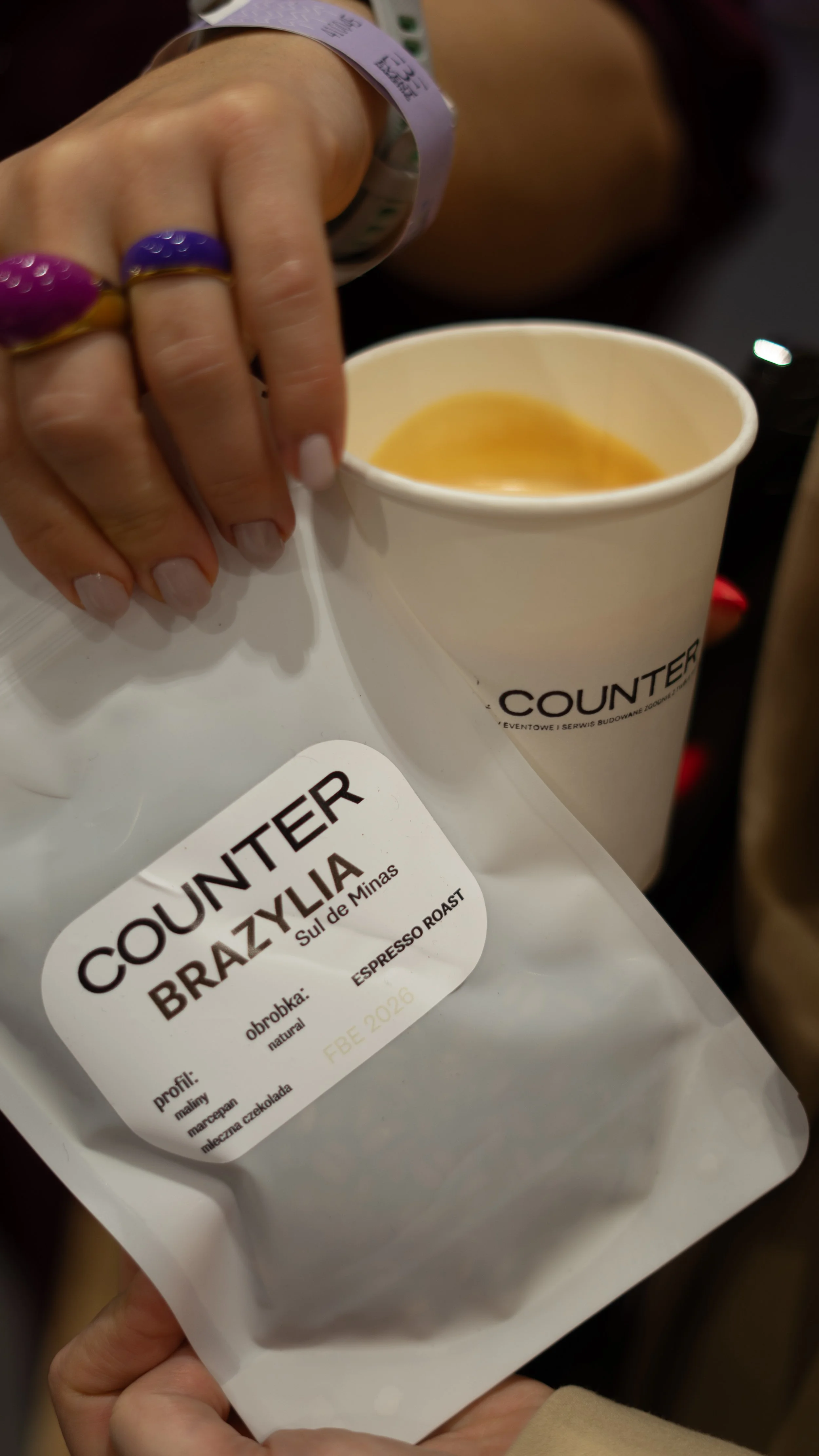 Person holding white coffee cup with coffee inside, labeled 'Counter,' with another hand holding a white package labeled 'Counter Brazylia Sul de Minas,' featuring details about the coffee's profile, processing method, and roast level.