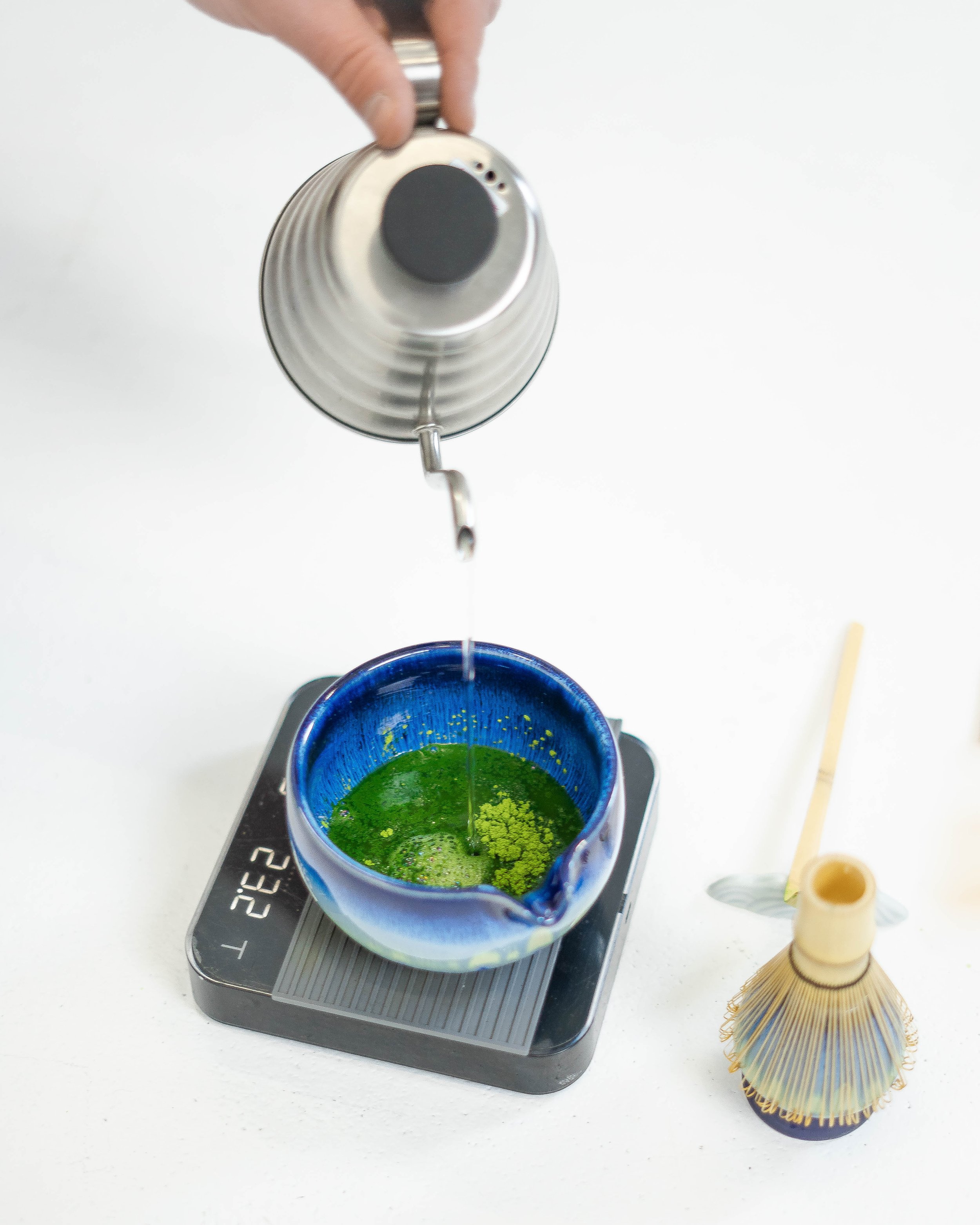 A hand pours hot water into a blue and green matcha tea bowl placed on a scale, with a bamboo whisk nearby.