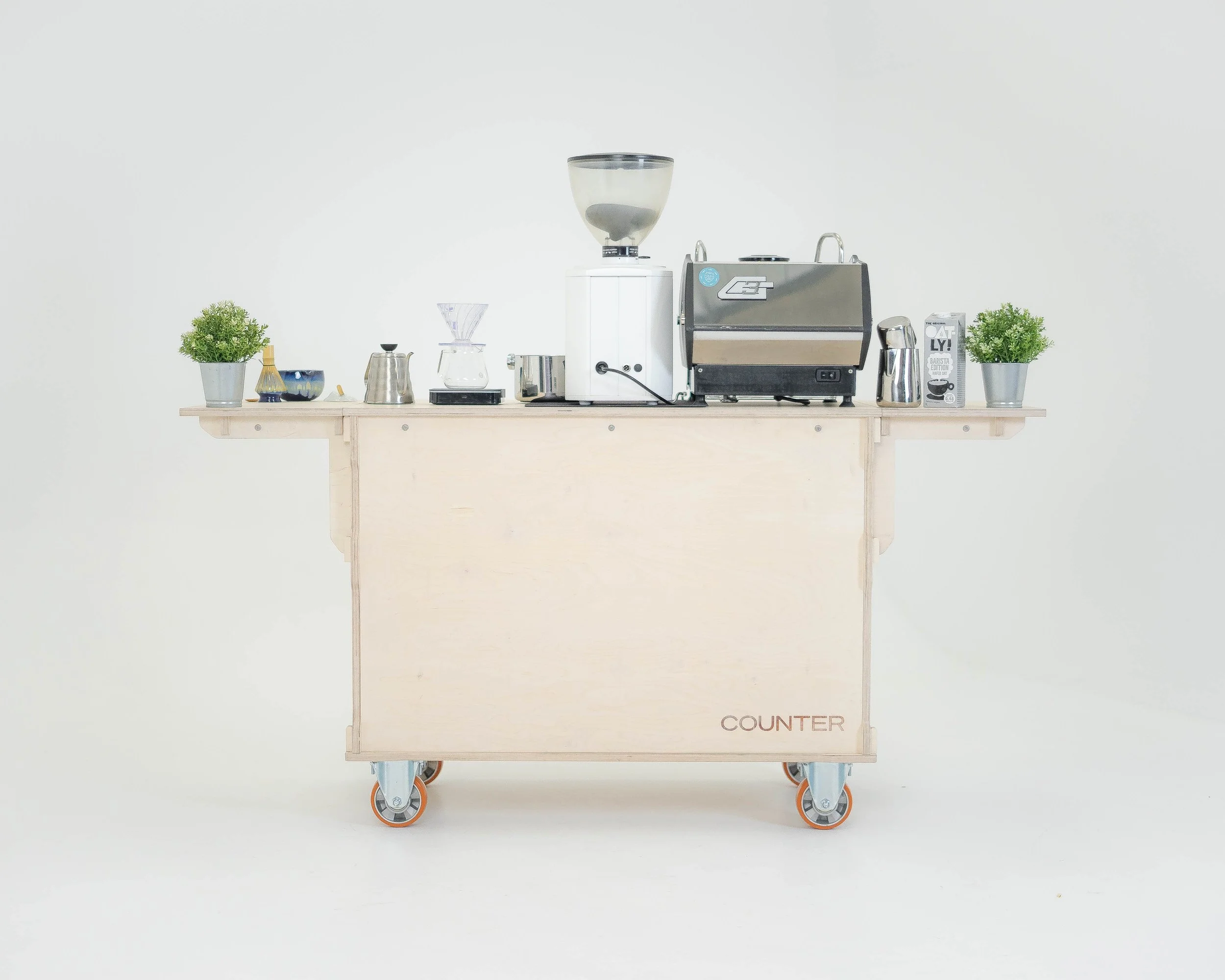 Coffee shop counter with various coffee-making equipment and potted plants on a wooden cart on wheels.