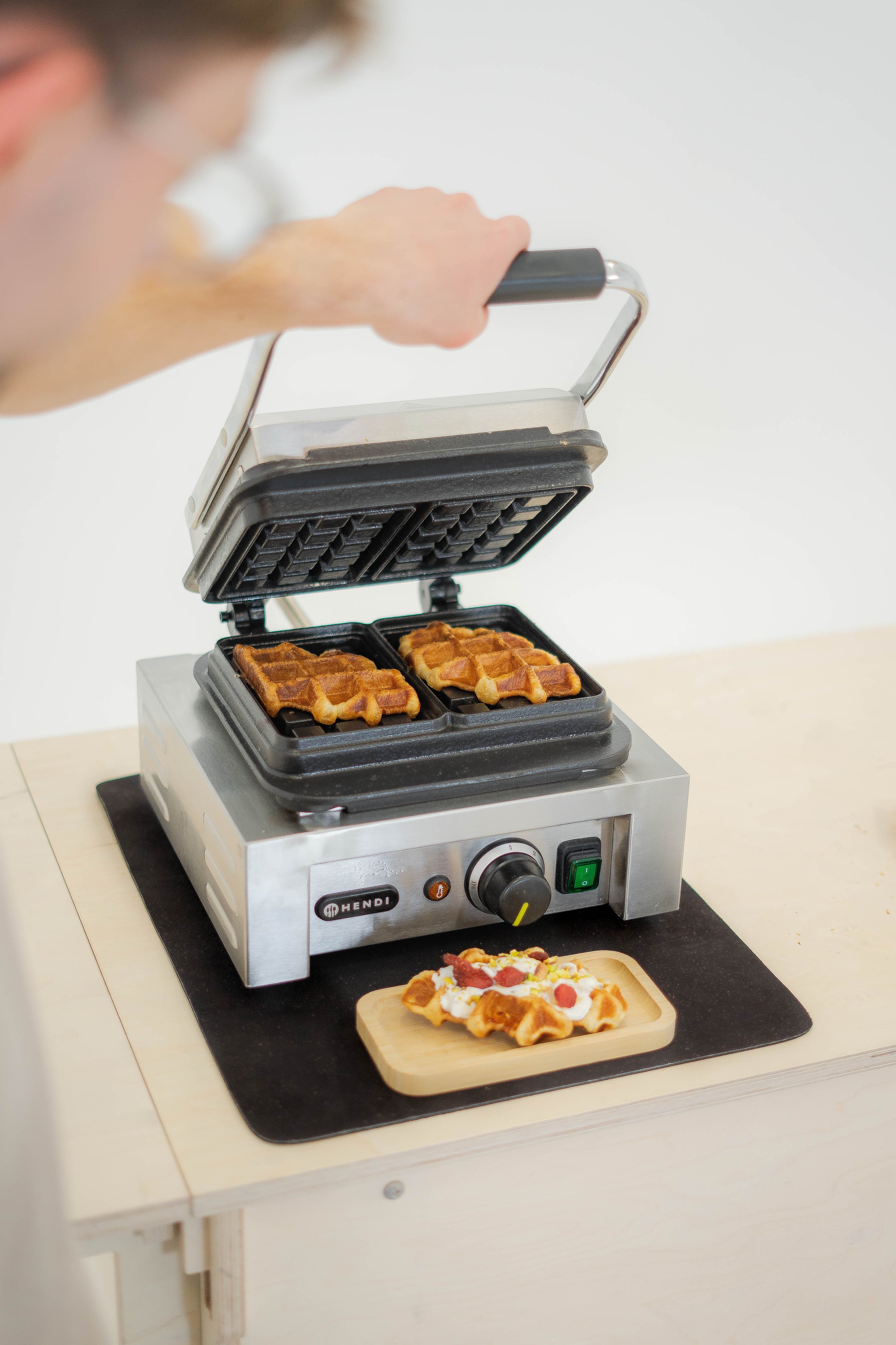 A person using a waffle maker to prepare waffles, with a plate of waffles topped with strawberries and whipped cream on the table.