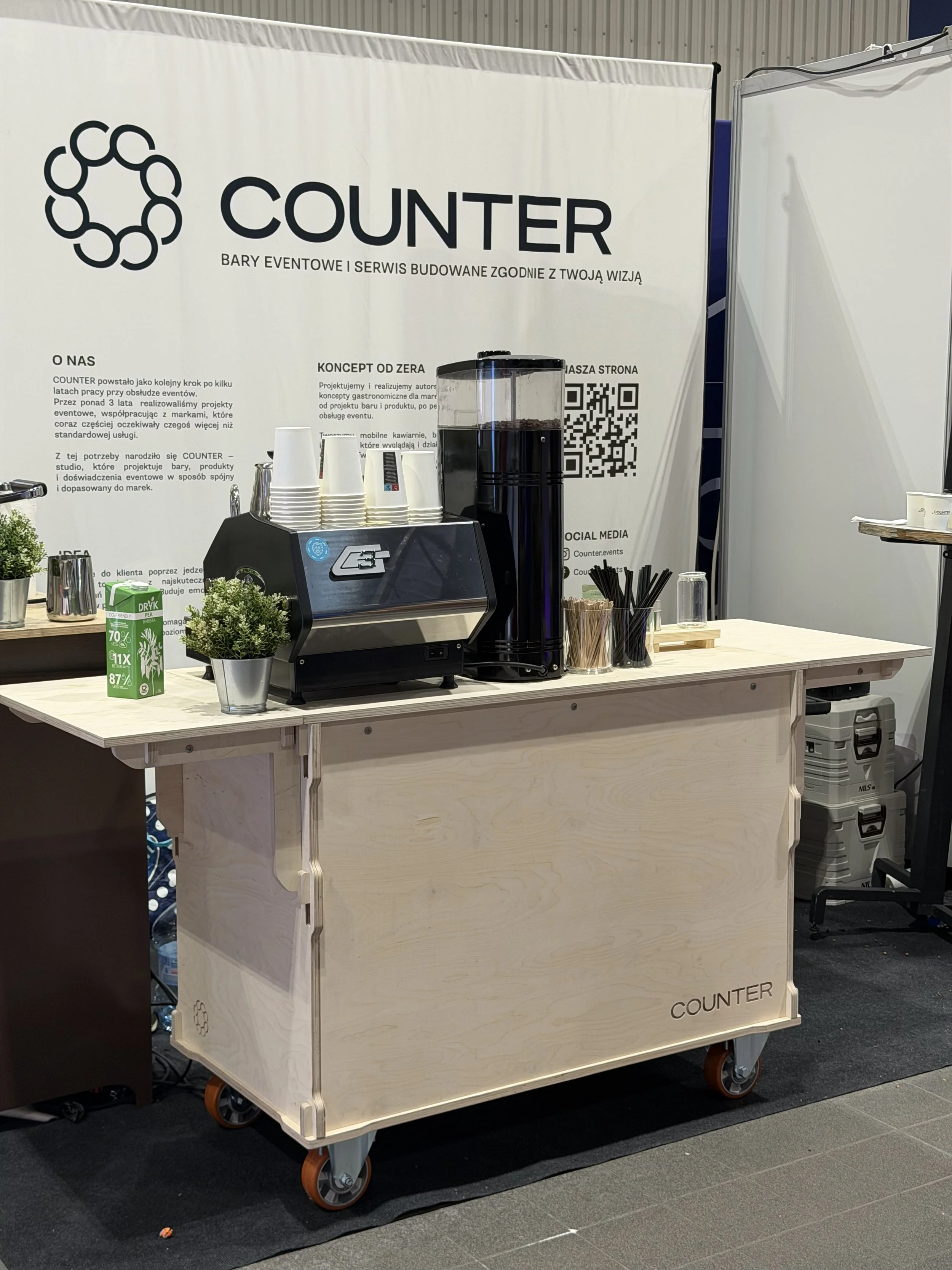 A mobile coffee station with a coffee machine, cups, and decorative plants in front of a banner for Counter event services.
