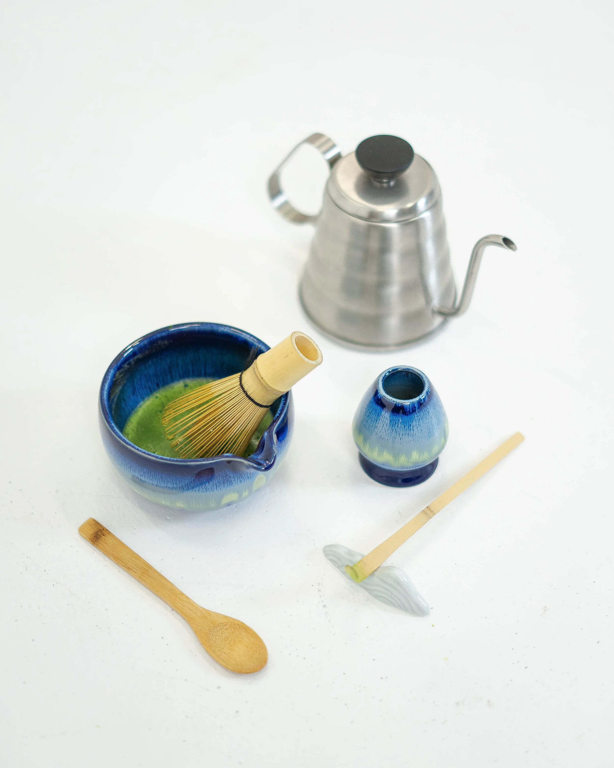 Tea set including a blue and green ceramic bowl, a small matching ceramic vase, a bamboo whisk in the bowl, a wooden spoon, a bamboo scoop, and a metal gooseneck kettle on a white background.