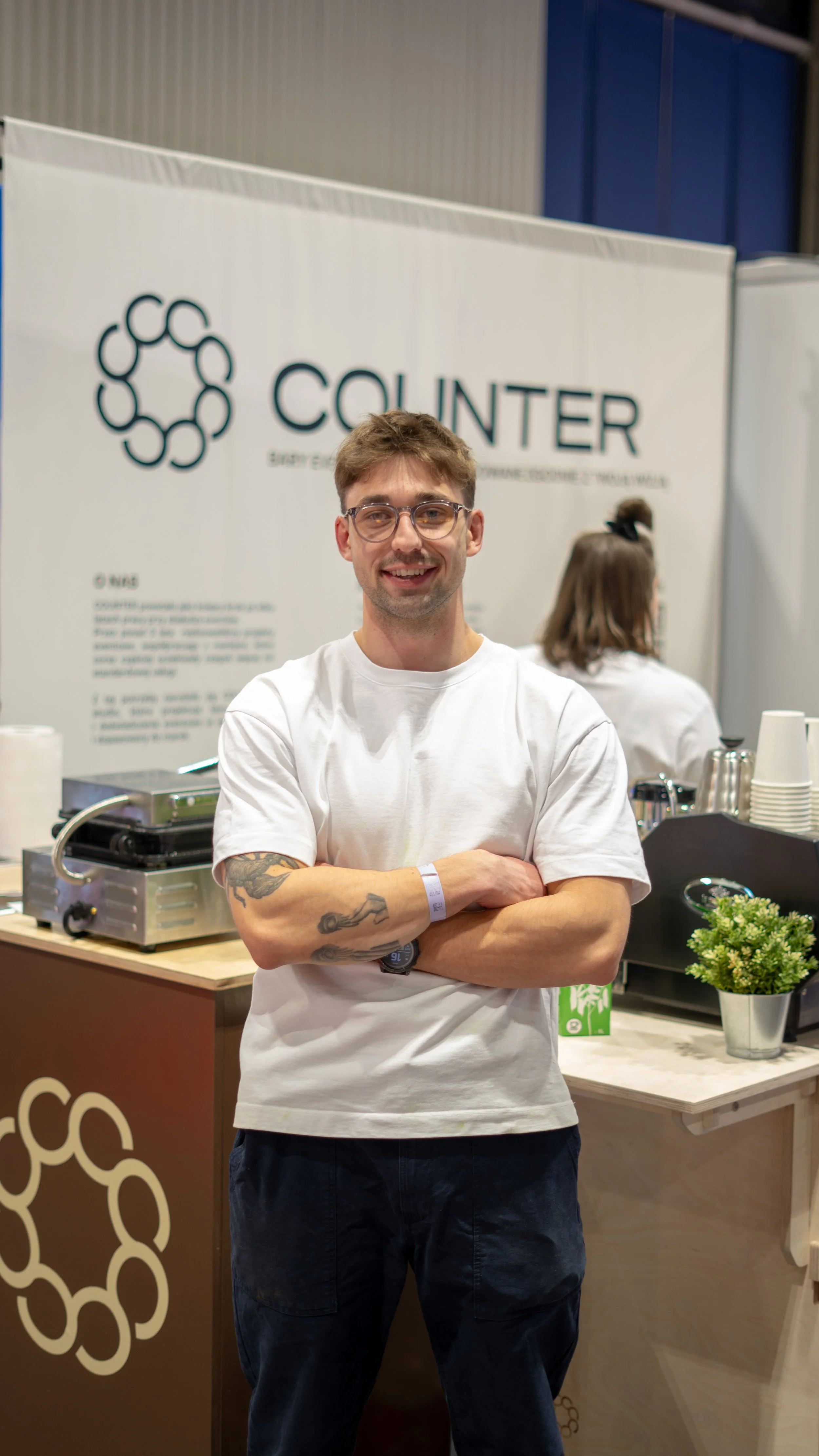 A man with glasses, tattoos on his arms, wearing a white T-shirt and a watch, standing with his arms crossed in front of a coffee stand with a sign that says 'COUNTER' with a logo of interconnected circles.