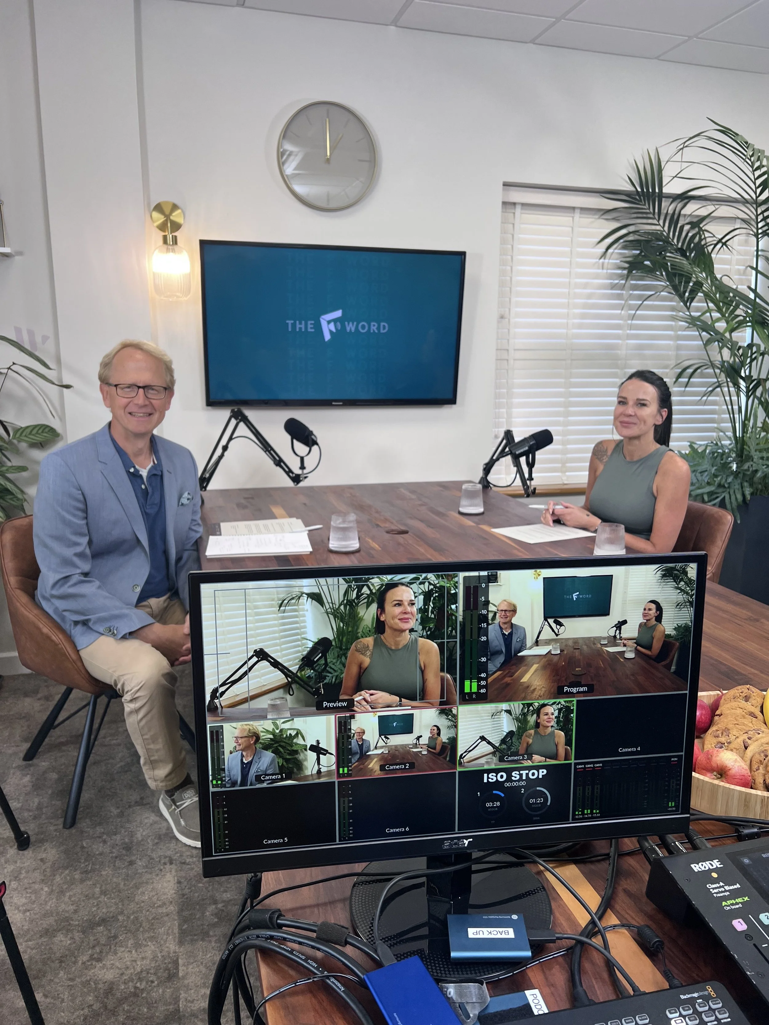 Two people are sitting at a table in a recording studio or office, with video cameras and equipment set up for a podcast or interview. There is a large monitor displaying the program logo, a clock, and some houseplants on the wall.