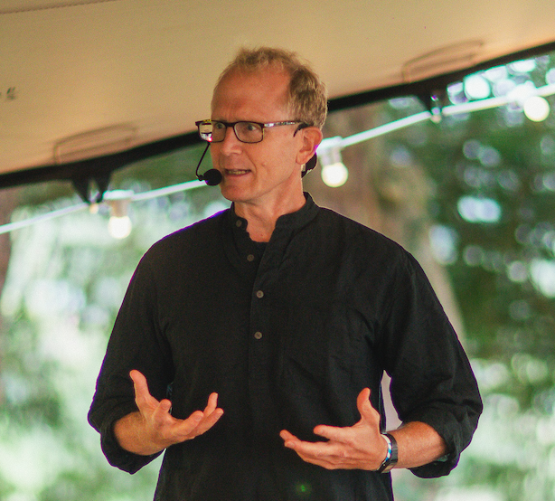 A man wearing glasses and a black shirt giving a presentation outdoors, with a headset microphone and gesturing with his hands.