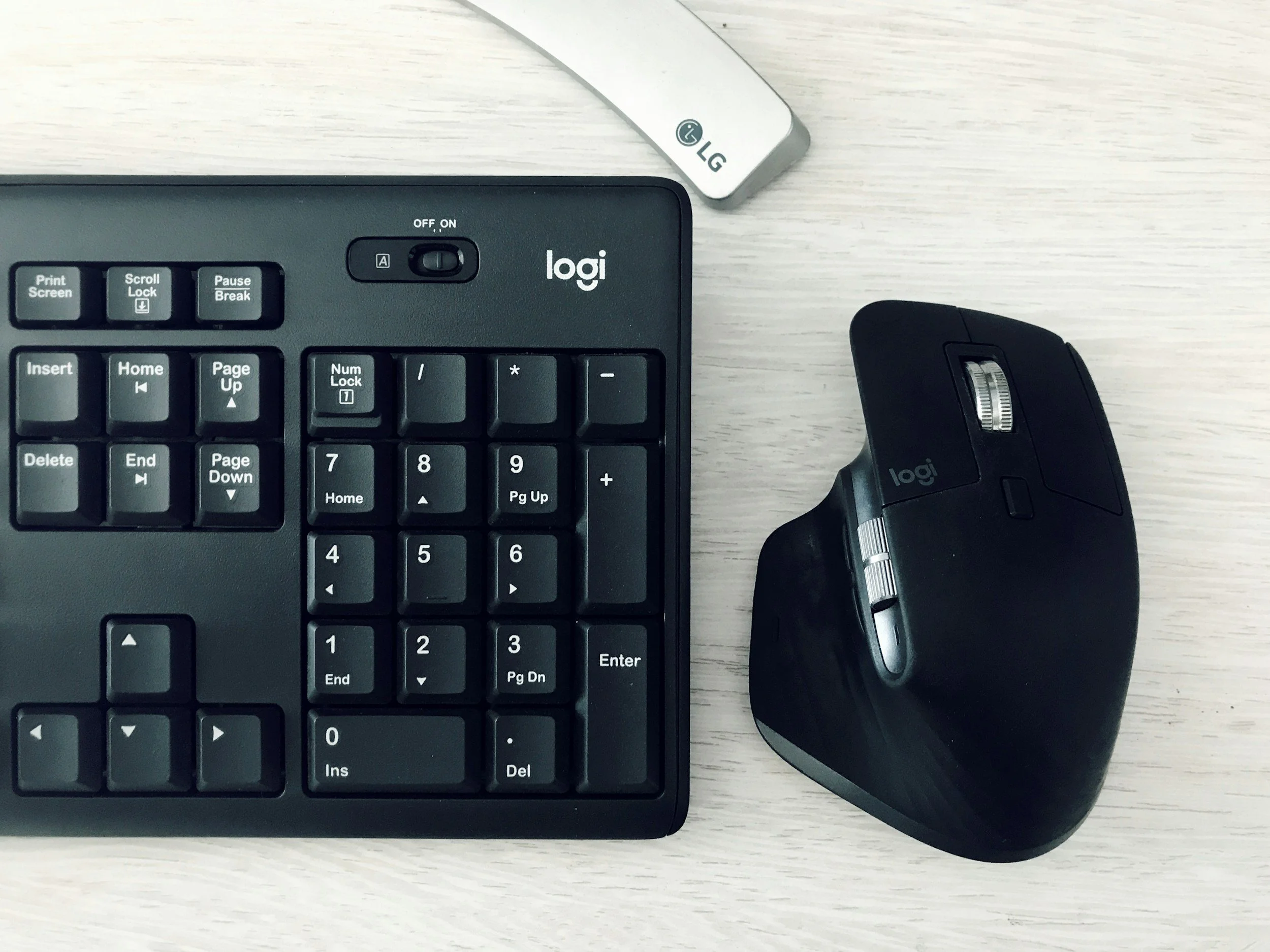 Close-up of a black computer keyboard with numeric keypad, a black Logitech mouse, a portion of a silver LG monitor stand, and a light-colored wooden desk surface.