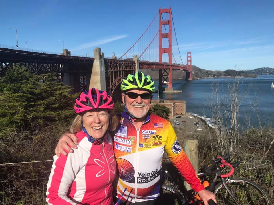 A smiling couple in cycling gear and helmets, standing in front of the Golden Gate Bridge in San Francisco, California.