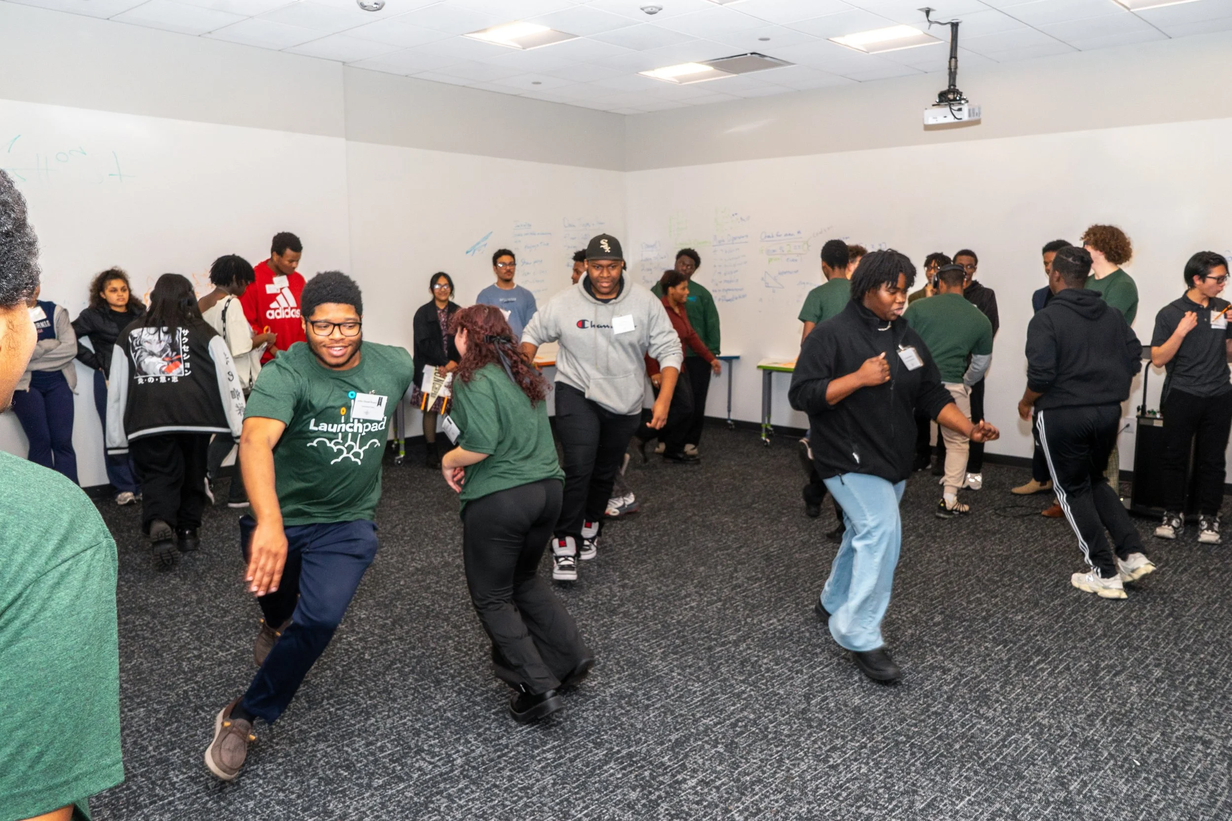 A group of young people dancing and socializing in a room with white walls and a dark carpeted floor, some standing against the wall, while others dance in the center.