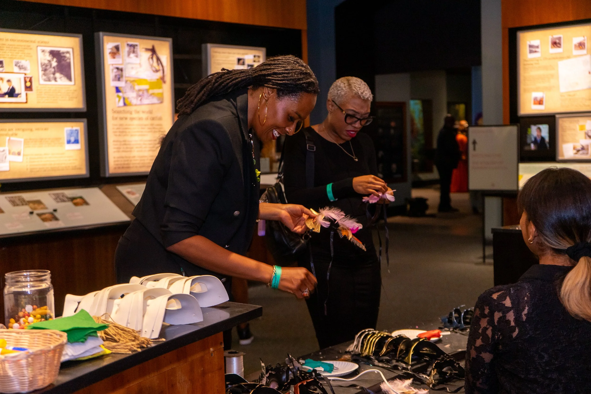 Two women are shopping for accessories at a booth, with one looking at feathered headbands and jewelry, while a third woman behind the table is assisting. The background shows exhibit displays and informational panels.