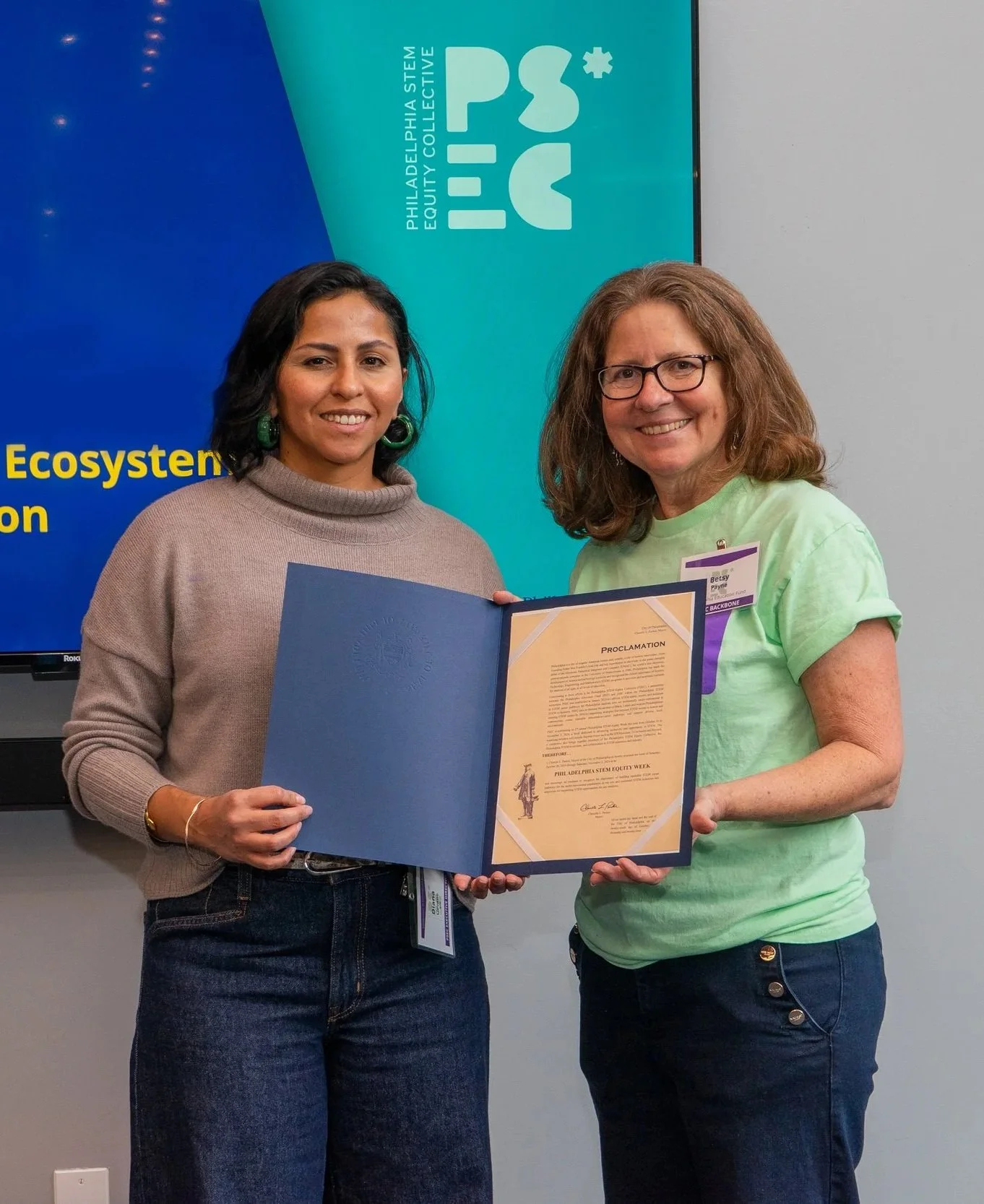 Two women smiling and holding a certificate, standing in front of a blue and teal banner with the logos of Philadelphia STEM Equity Collective.