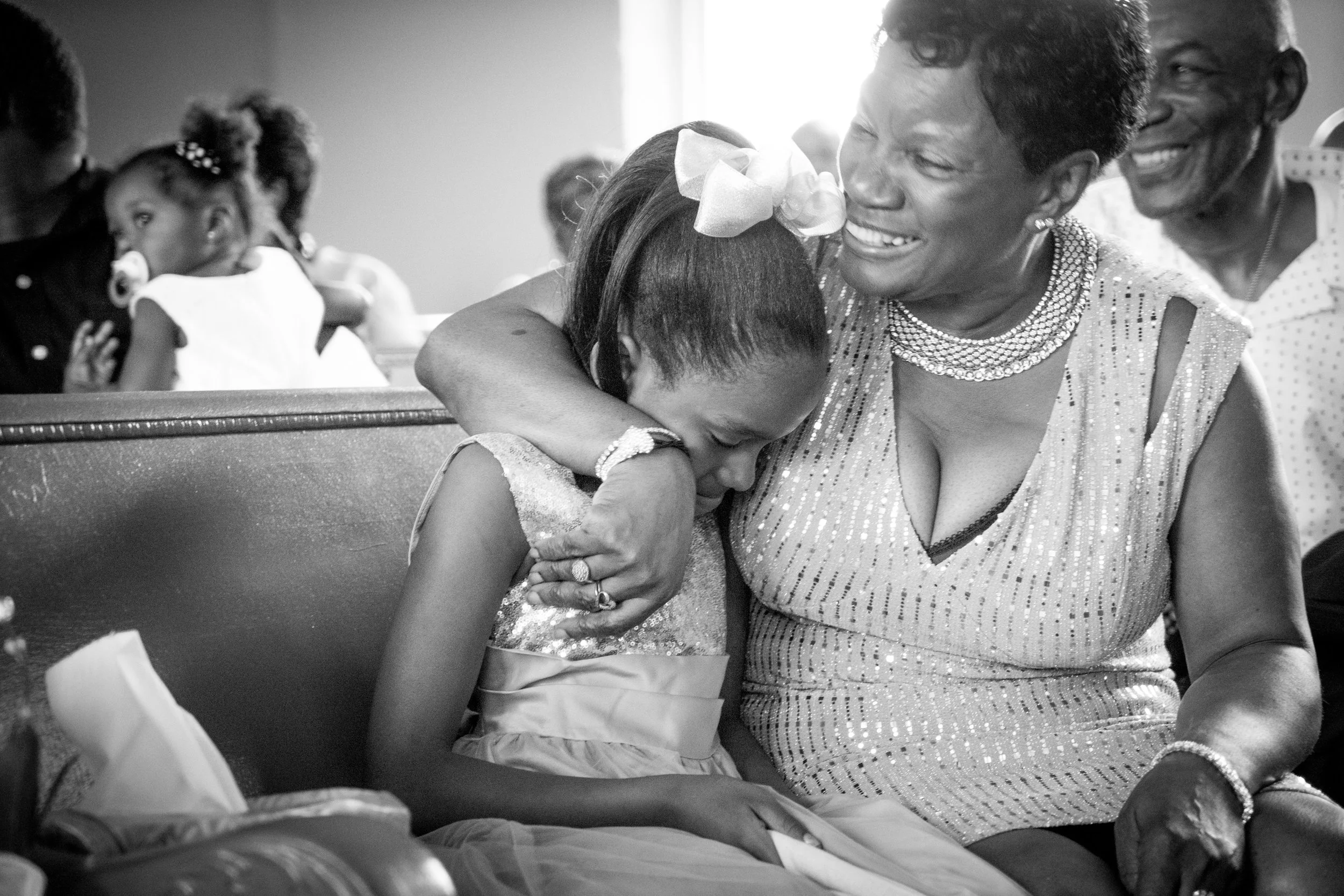 A black-and-white photo of a woman and a girl sharing a hug, both smiling and showing emotion. In the background, other people are visible, including a girl with a pacifier and an adult woman.