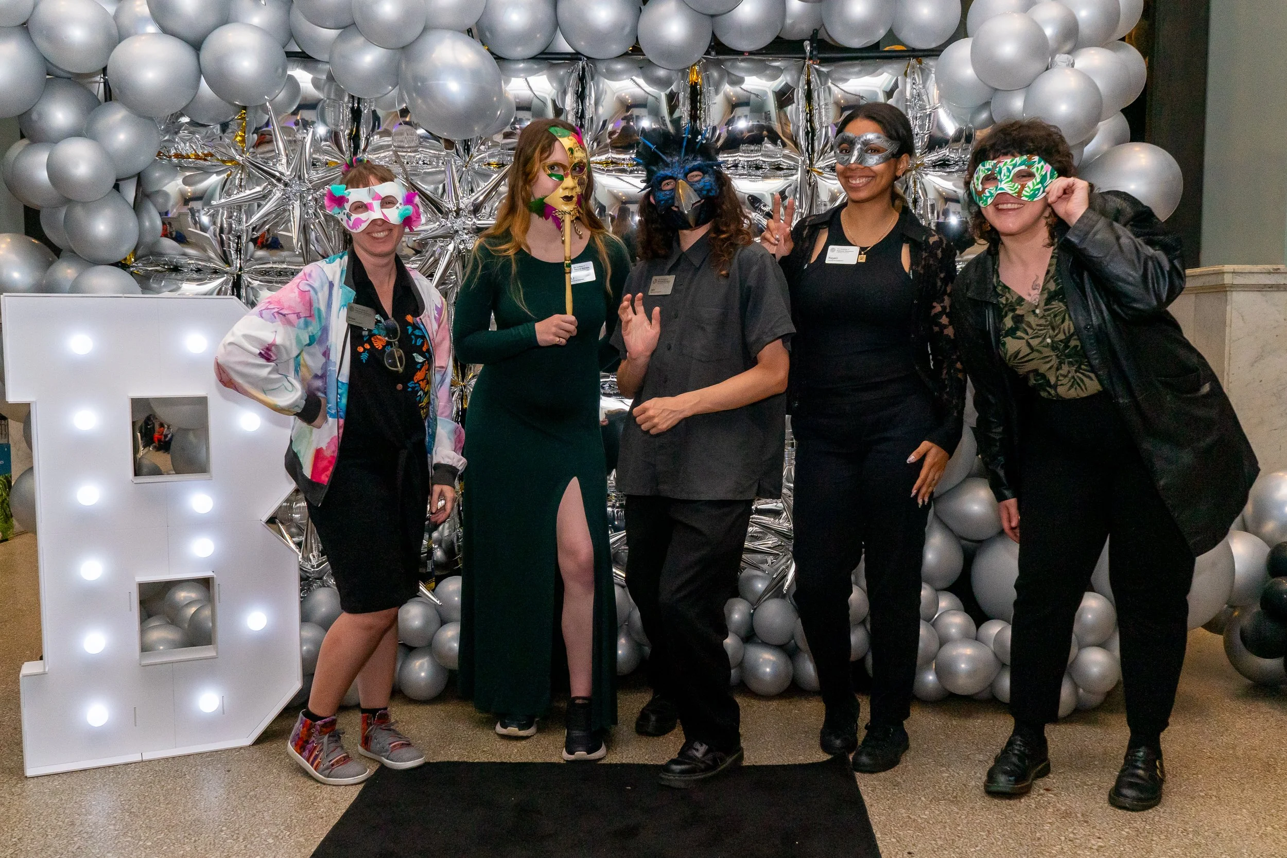 Five women wearing decorative masks and holding props at a festive event, standing in front of a backdrop made of silver balloons and a large illuminated letter 'B'.