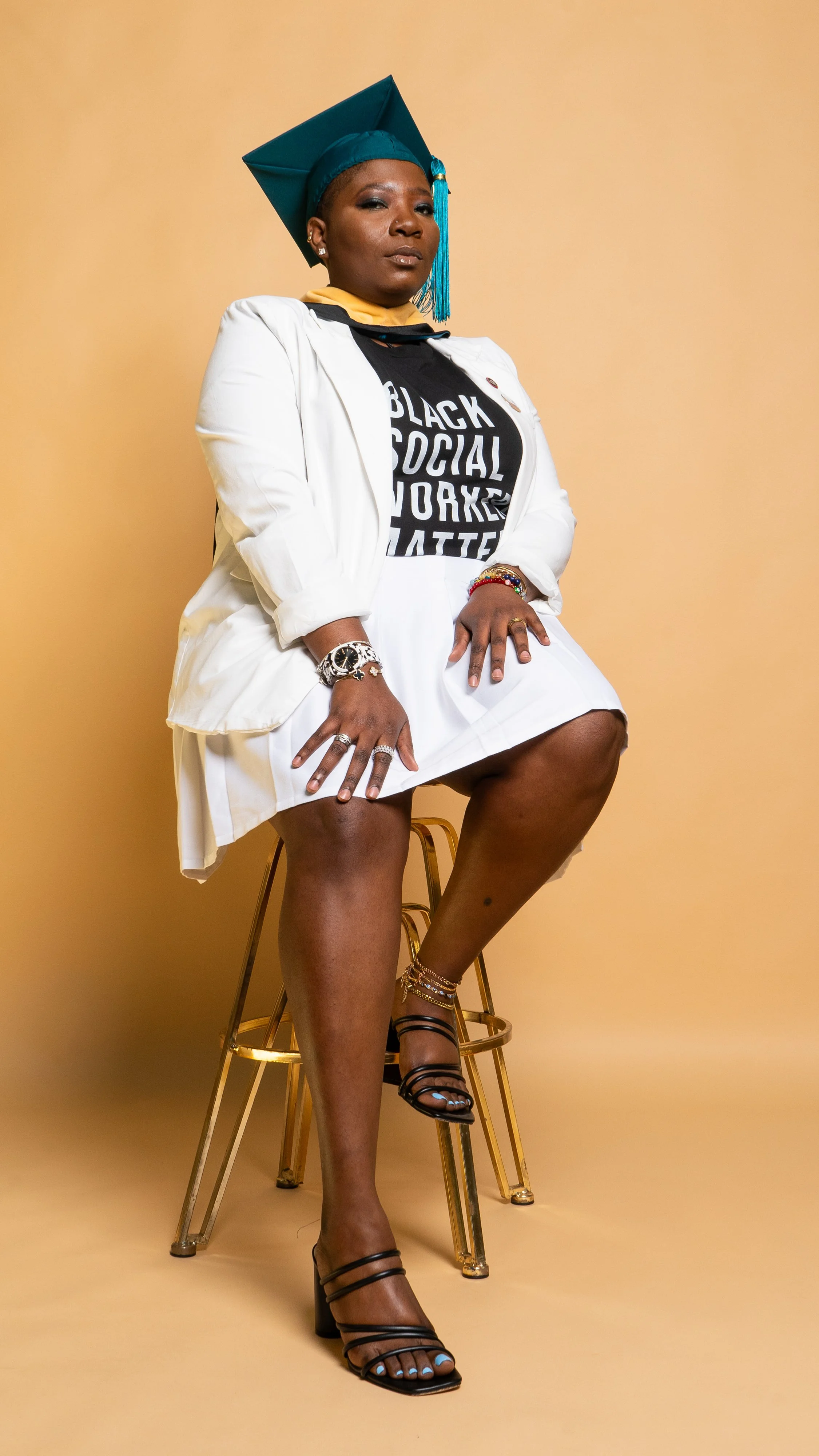 A woman wearing a graduation cap and gown sits on a gold chair against a beige background. She is dressed in a black T-shirt with the words 'Black Social Worker' printed on it, a white skirt, and black strappy high heels. She has jewelry on her wrist