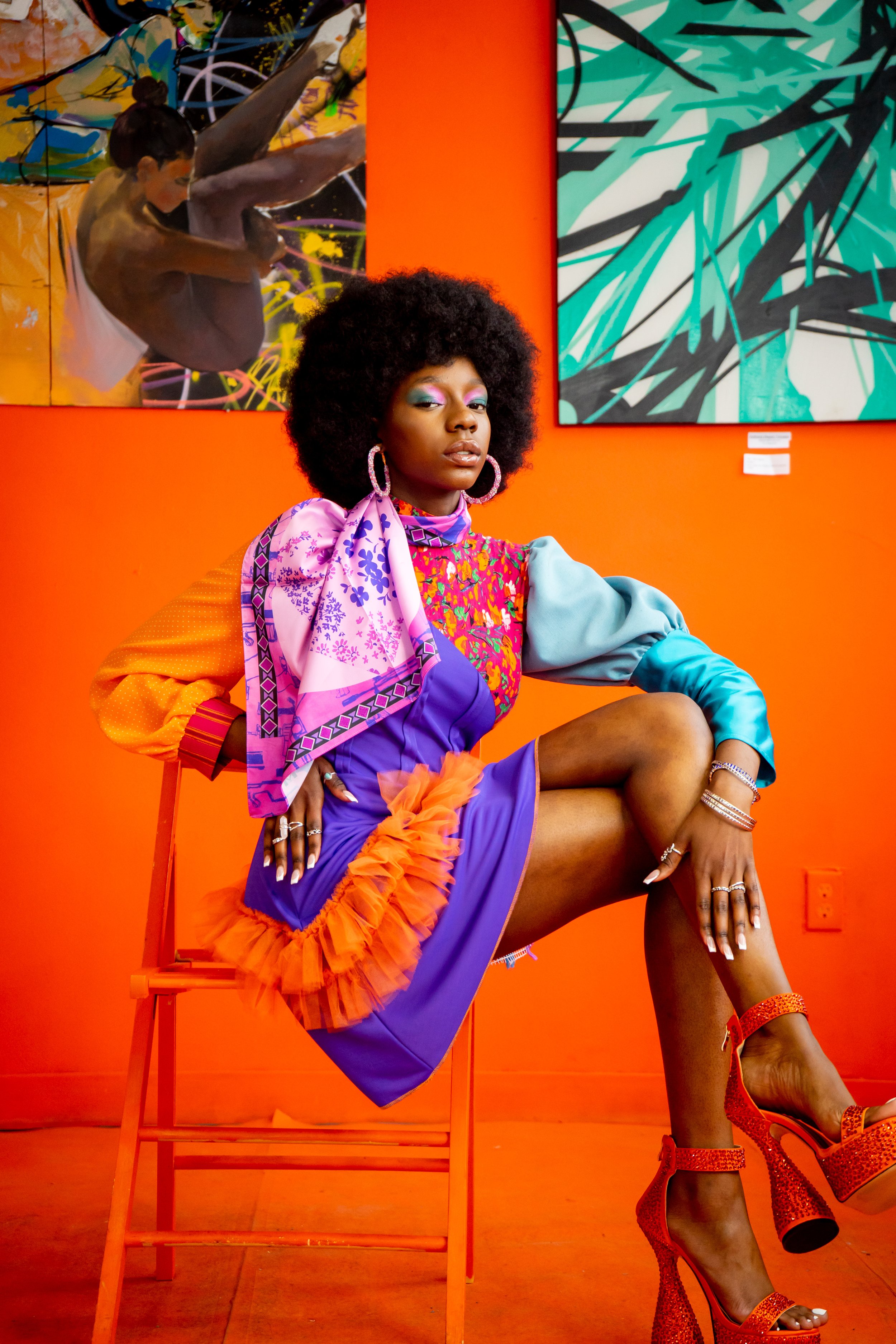 A woman sitting on an orange chair against an orange wall with colorful abstract paintings. She is wearing vibrant, eclectic clothing with a ruffled orange and purple dress, large hoop earrings, and red high heels. She has a curly afro hairstyle and 