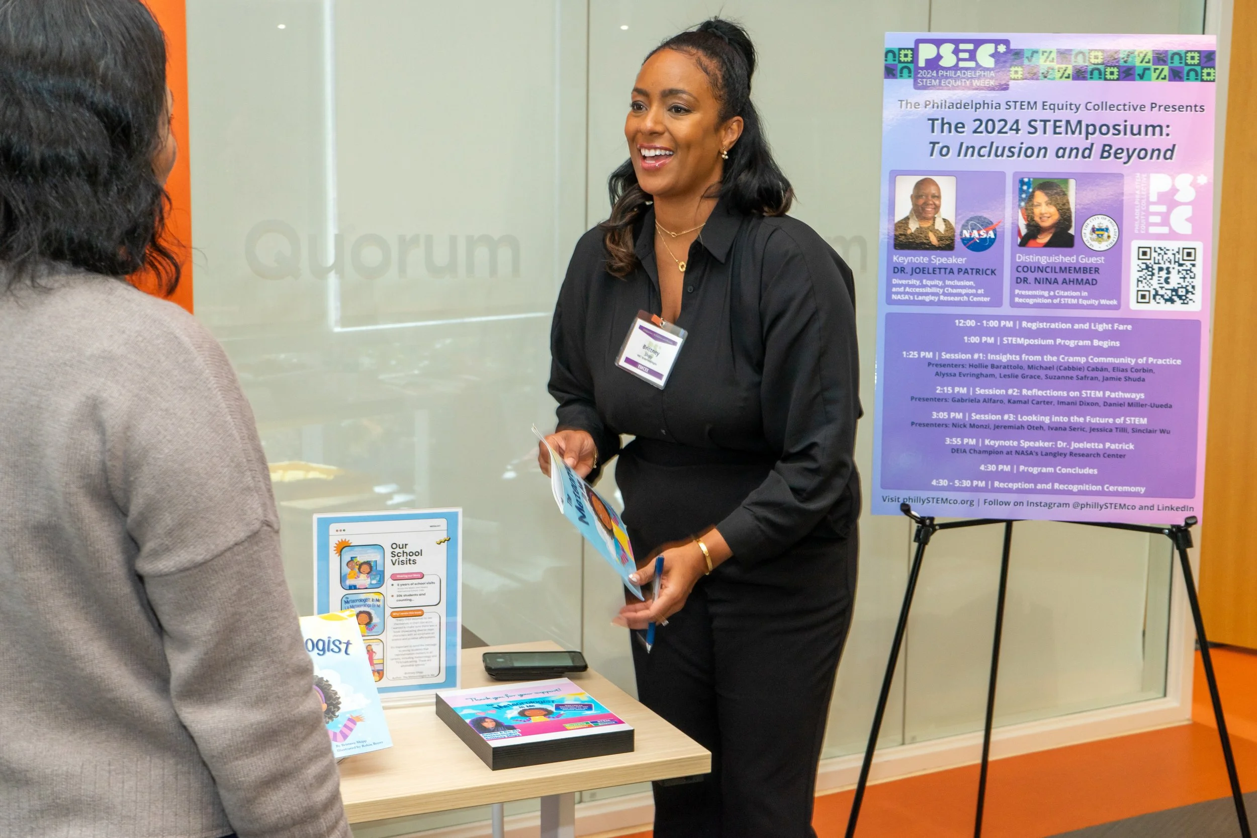A woman in black talking to another person at a conference registration table. A large posterBoard displays the agenda for a STEM symposium behind them.