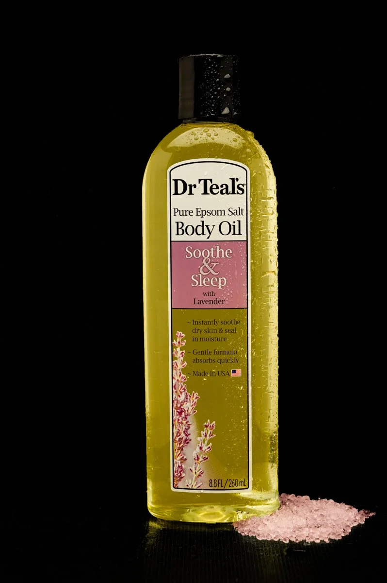 A bottle of Dr Teal's Soothe & Sleep Lavender Body Oil with pink bath salts spilled at the base, set against a black background.