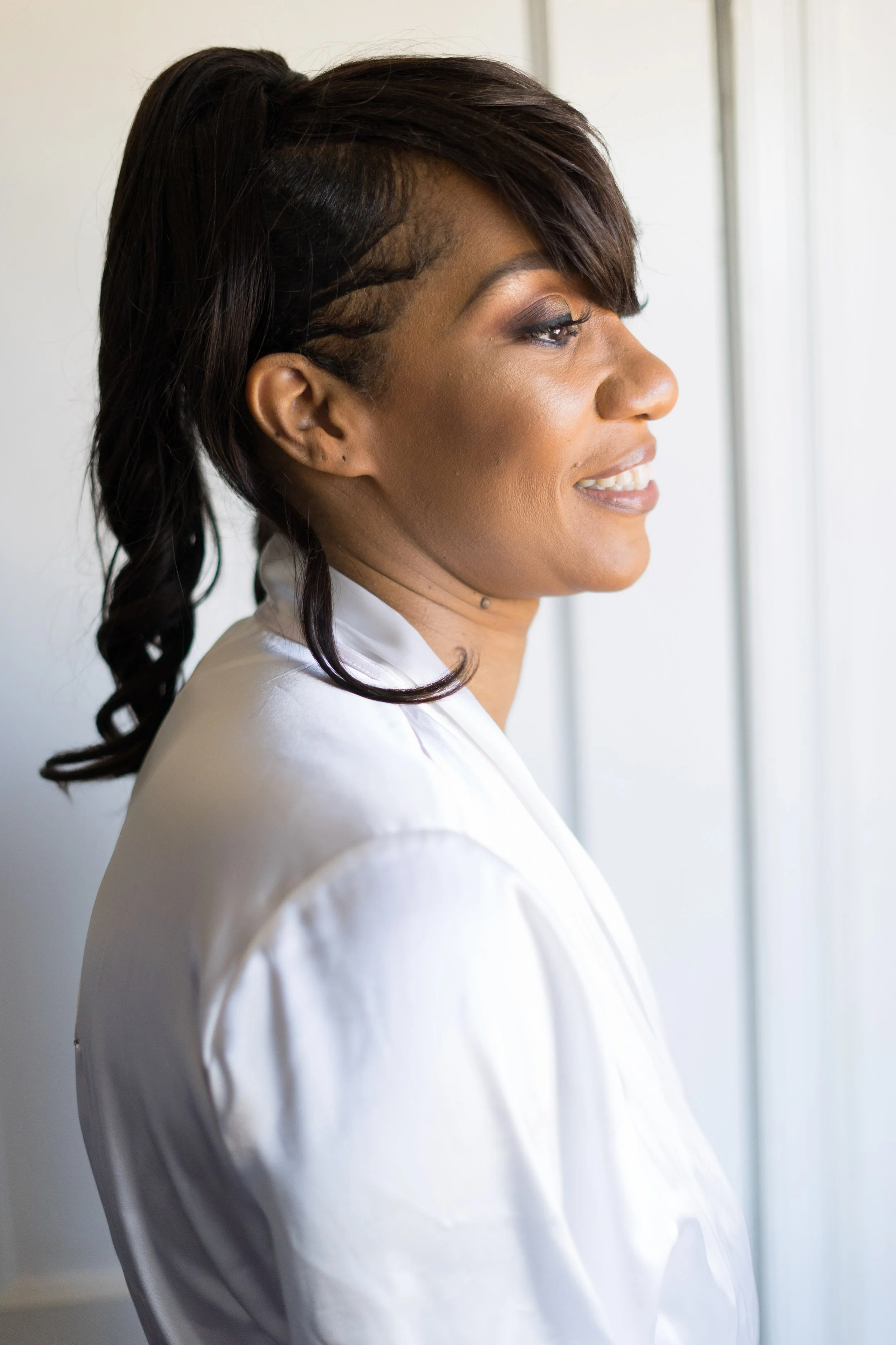 Side profile of an African American woman with styled hair, smiling, wearing a white satin robe, looking out a window.