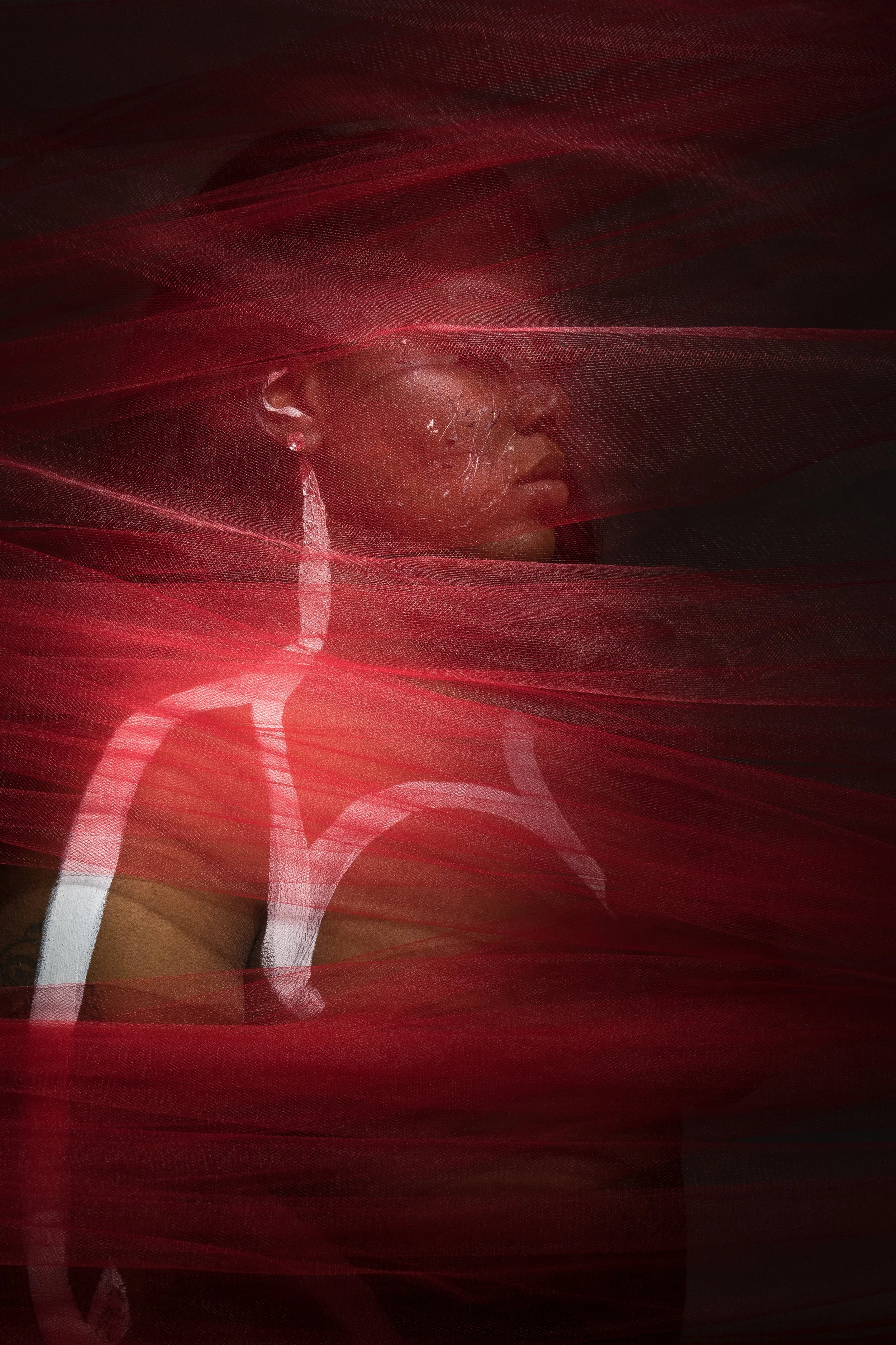 A woman with large earrings and a painted white abstract design on her shoulder, partially obscured by flowing red fabric or tulle.