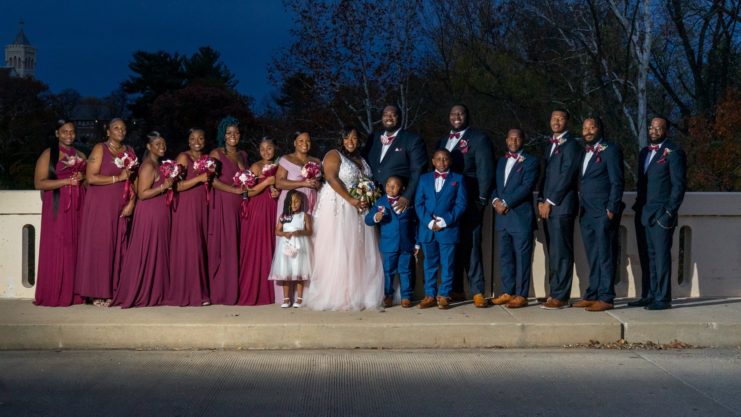 Group of people at a wedding, including the bride and groom, bridesmaids in burgundy dresses, groomsmen in navy suits with red bow ties, and children in formal attire, standing outdoors on a bridge during evening.