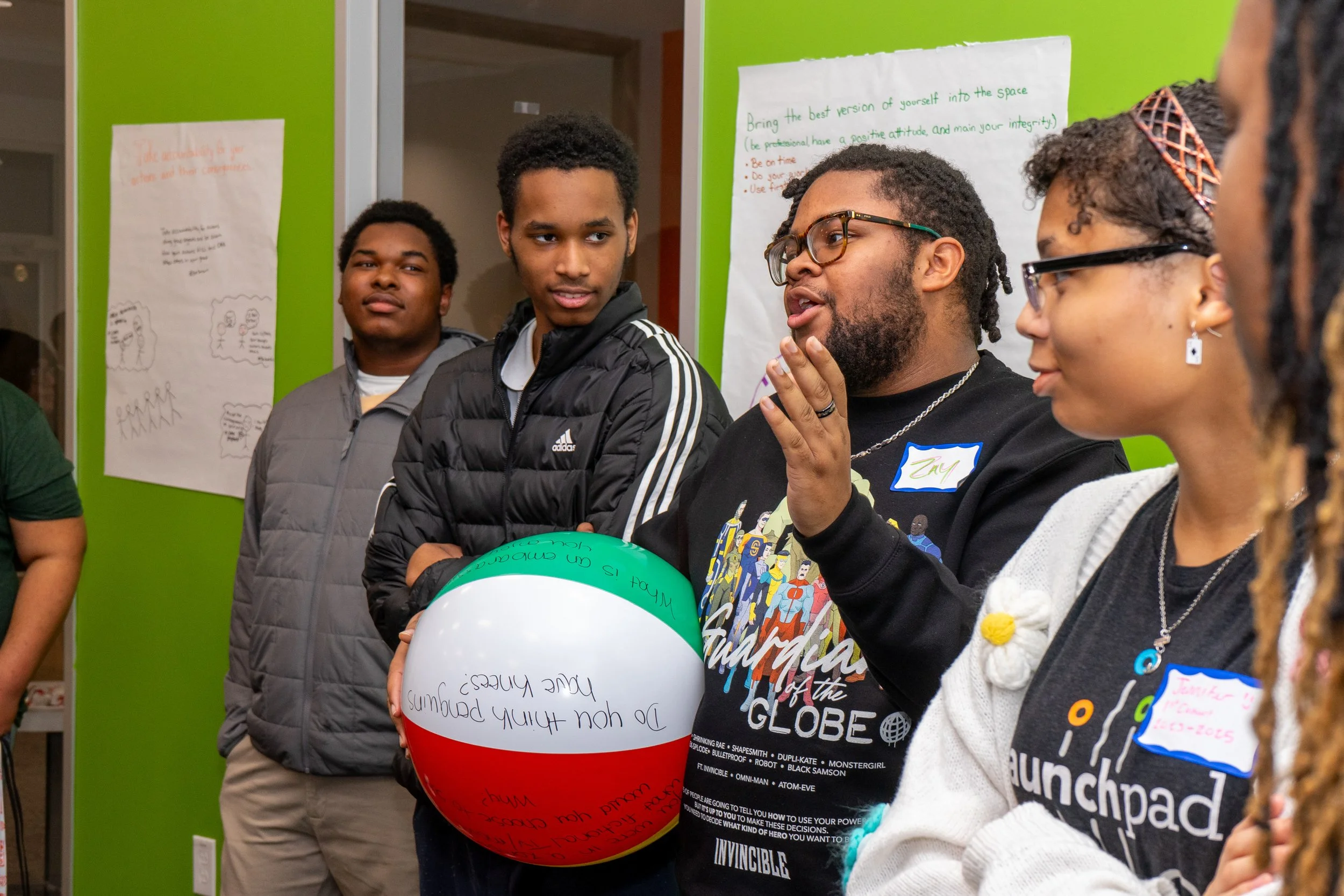 Group of young people standing in a room with green walls, listening to a man who is speaking. One person holds a large beach ball decorated like the Italian flag. The background has large sheets of paper with handwritten notes.