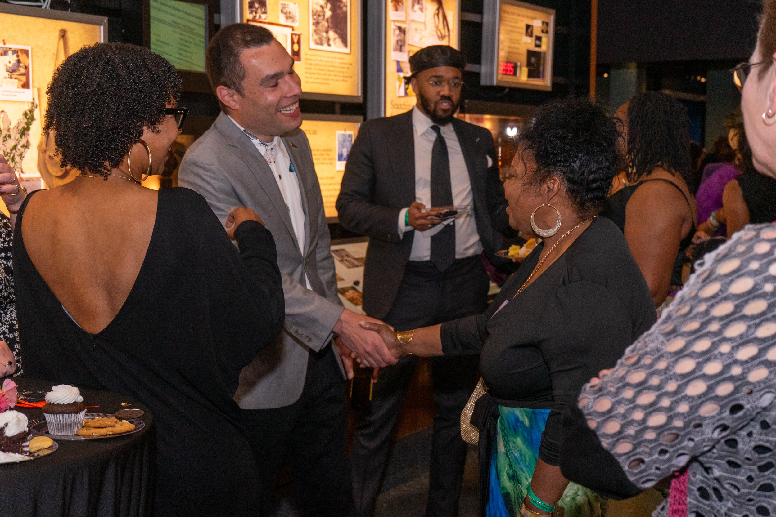 People at a social event engage in conversation. A man in a gray suit shakes hands with a woman in a black dress. Others in the background are talking, some holding plates of food. There is a table with cupcakes and cookies on the left.