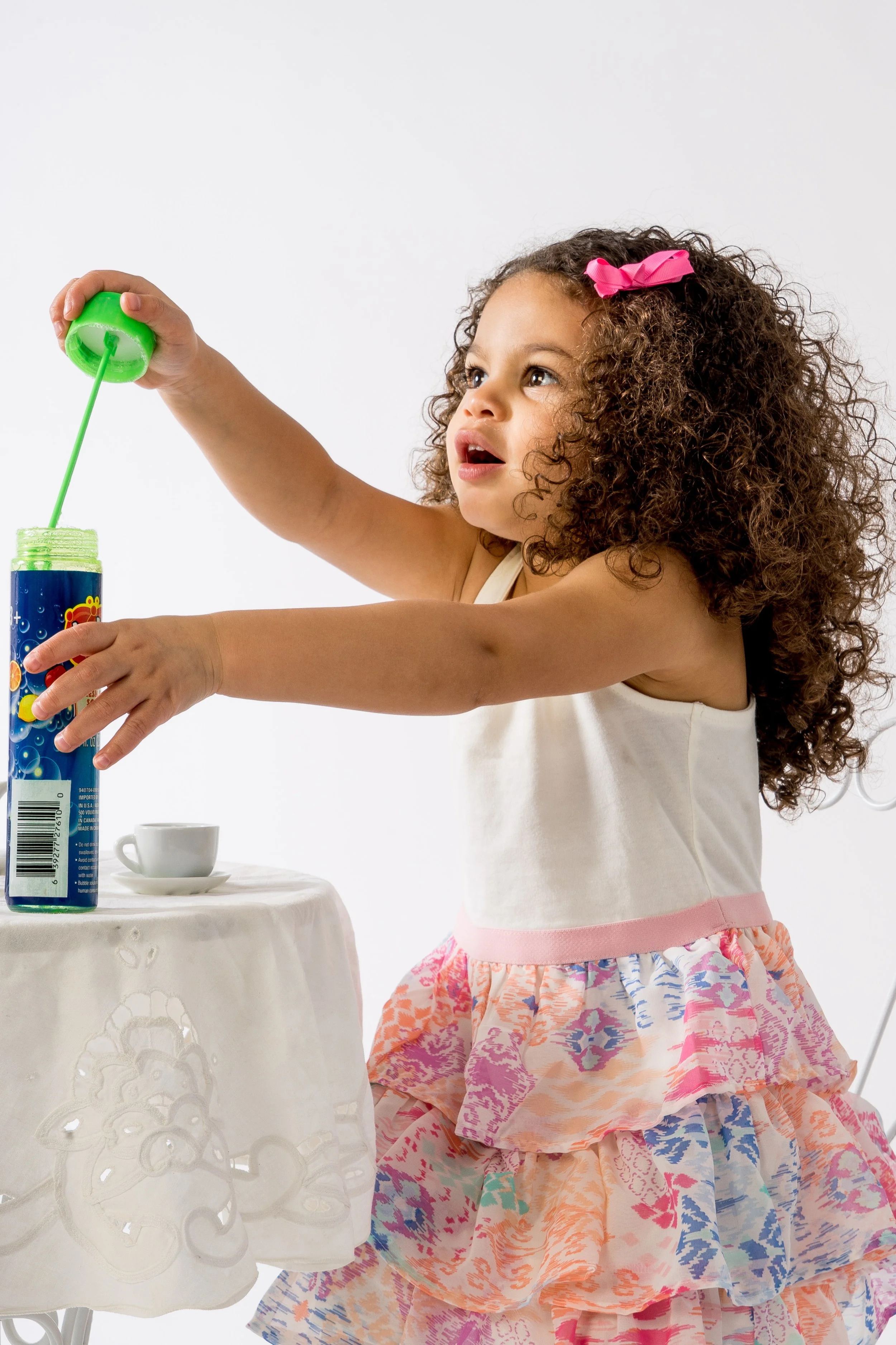 A young girl with curly hair and a pink bow, wearing a white top and a patterned skirt, is blowing bubbles from a bubble wand.