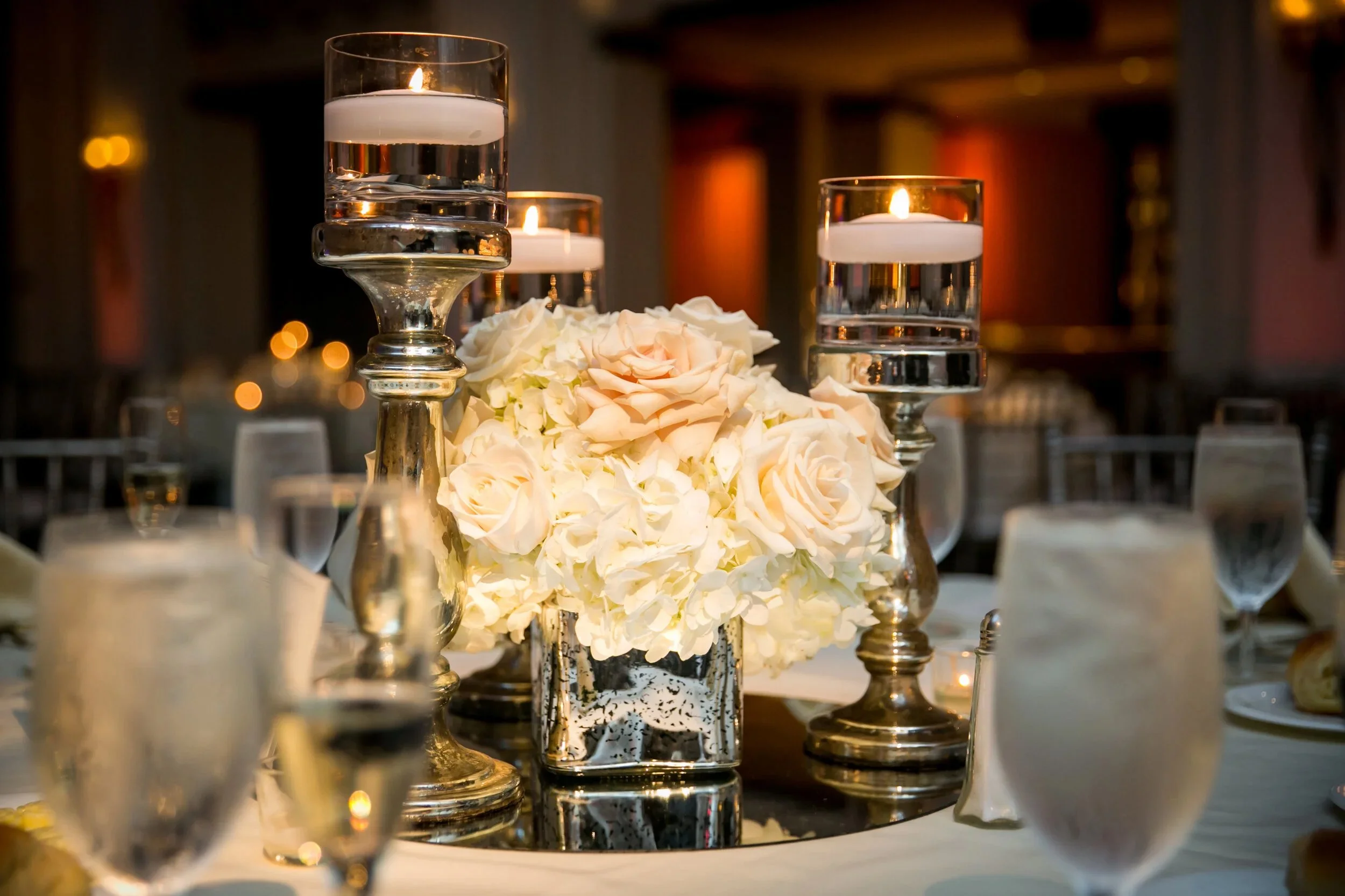 Elegant table centerpiece with white flowers, tall glass candle holders with floating candles, and glassware at a formal event.