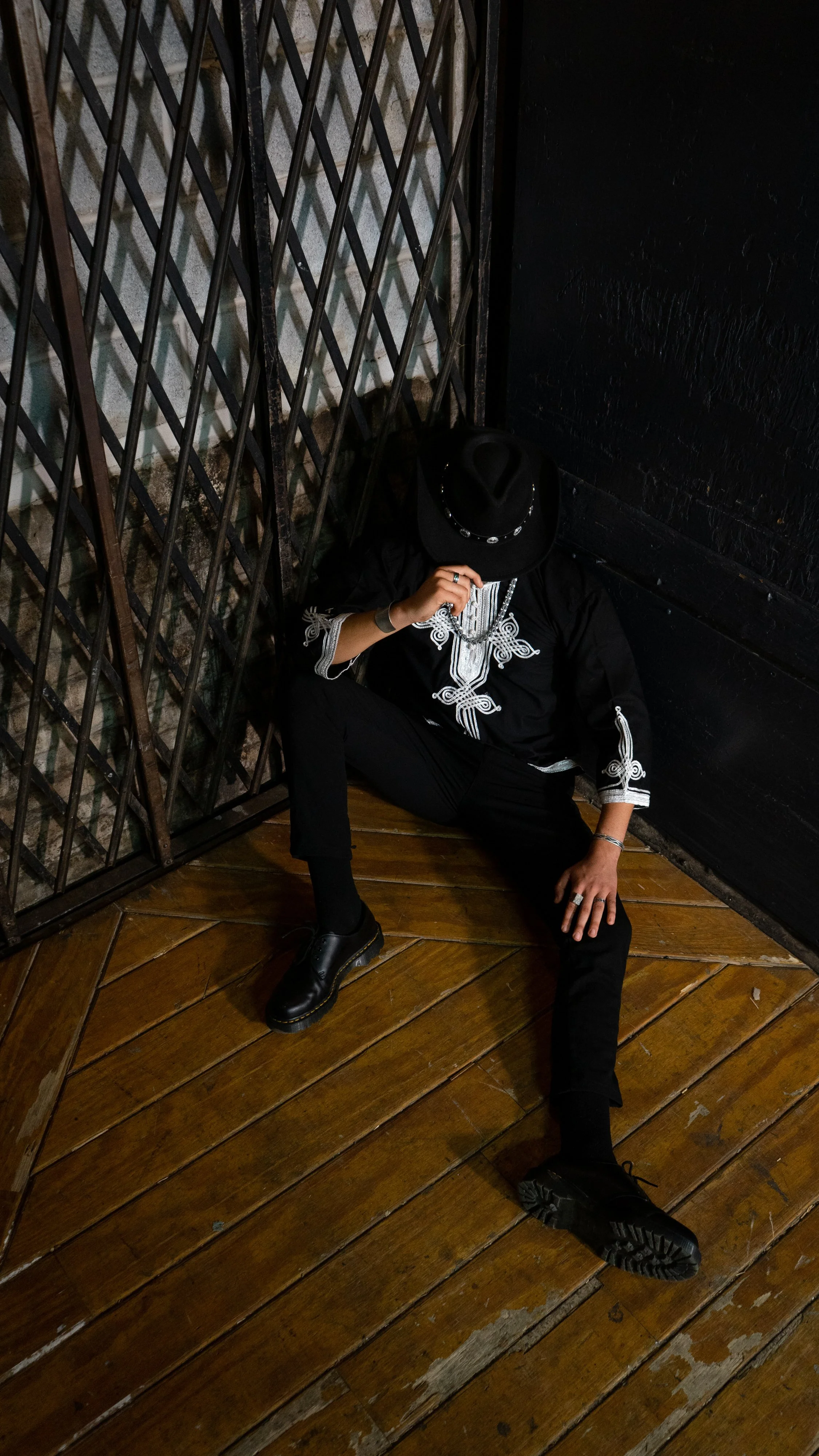 Person dressed in black with embroidered details, sitting on wooden floor with legs apart, wearing black shoes, black hat, and jewelry, leaning against a black wall with metal bars nearby, in an industrial or artistic space.