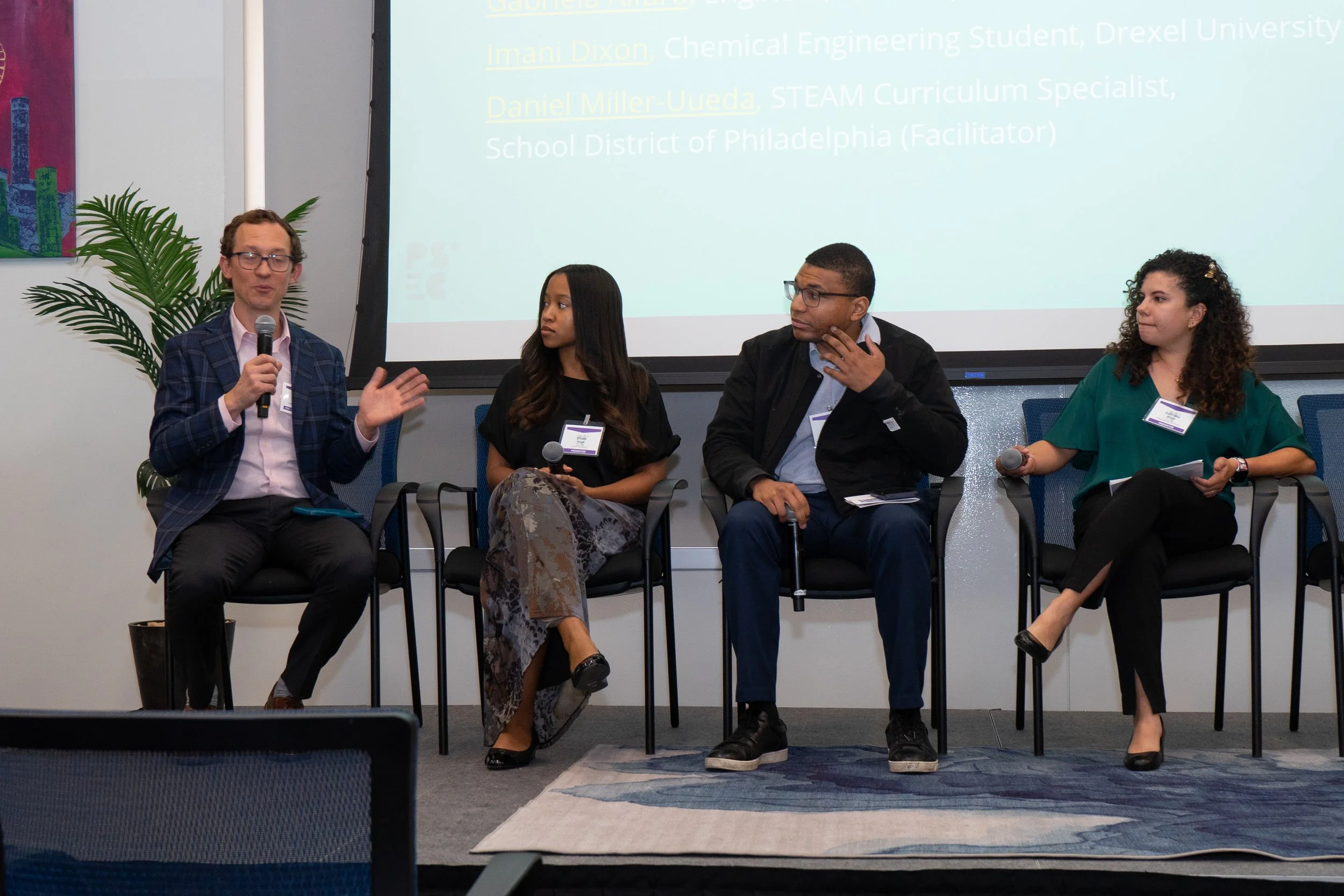 Panel discussion with four diverse individuals seated on stage, one speaking into a microphone, and a large screen behind displaying text with names and titles, in a conference room.