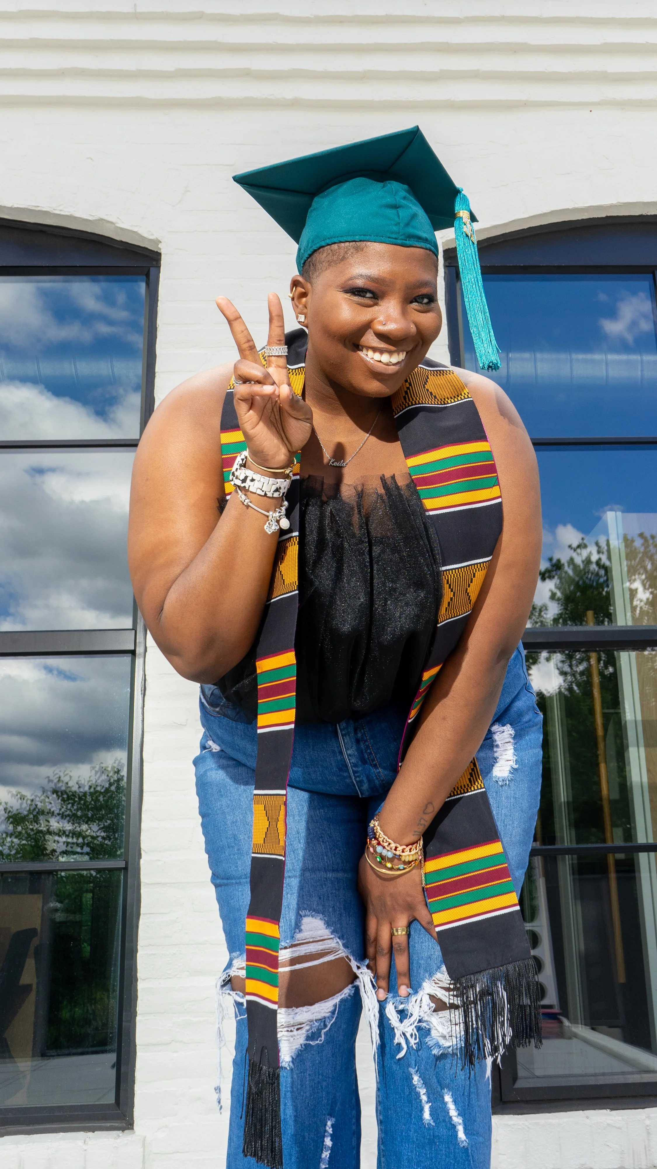A young woman in a teal graduation cap and colorful sash celebrating outside, smiling, making a peace sign with her right hand, and wearing ripped jeans, accessorized with bracelets and rings.