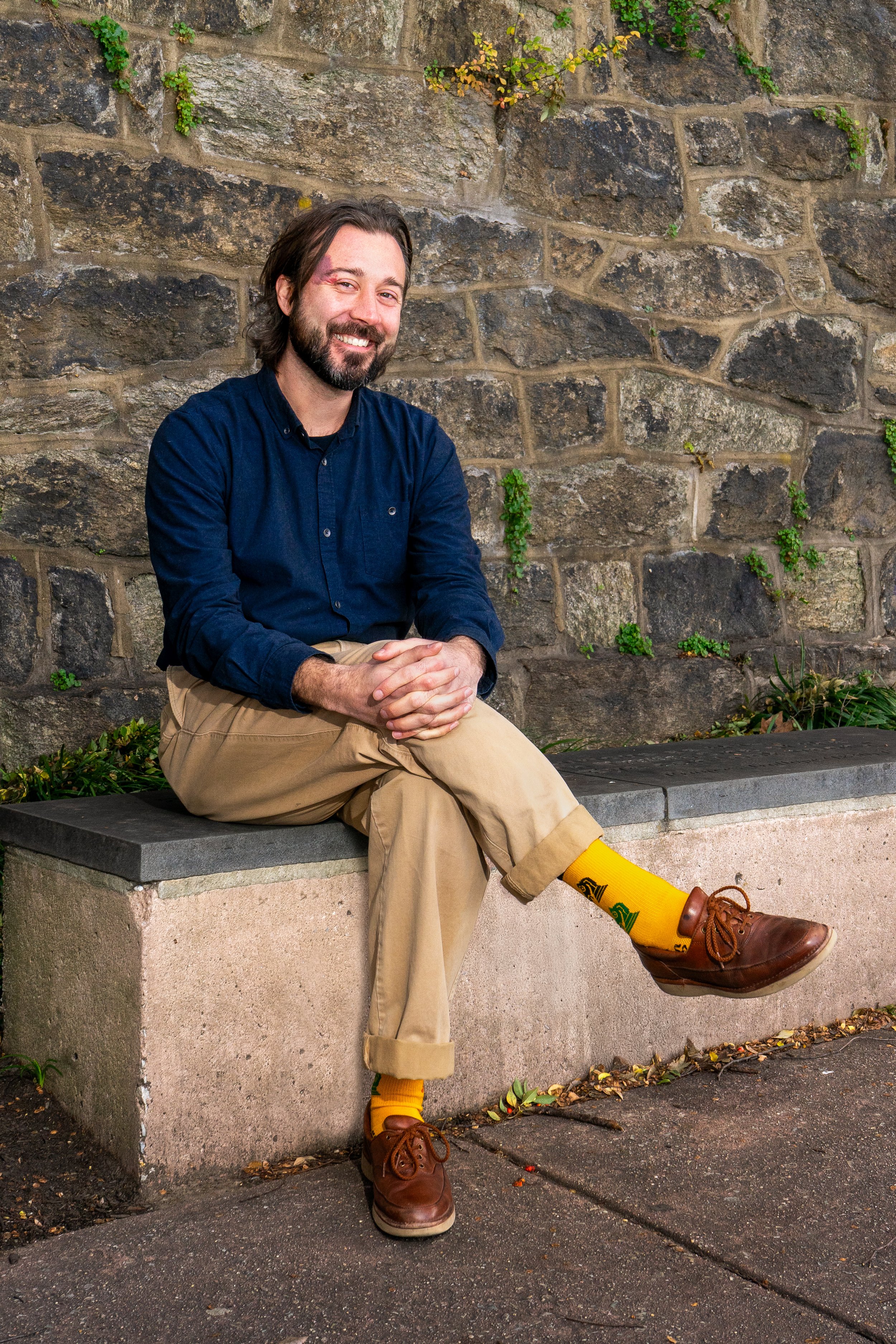 A man with dark hair and a beard sitting on a bench against a stone wall, smiling at the camera. He is wearing a navy blue shirt, khaki pants, yellow socks, and brown shoes.