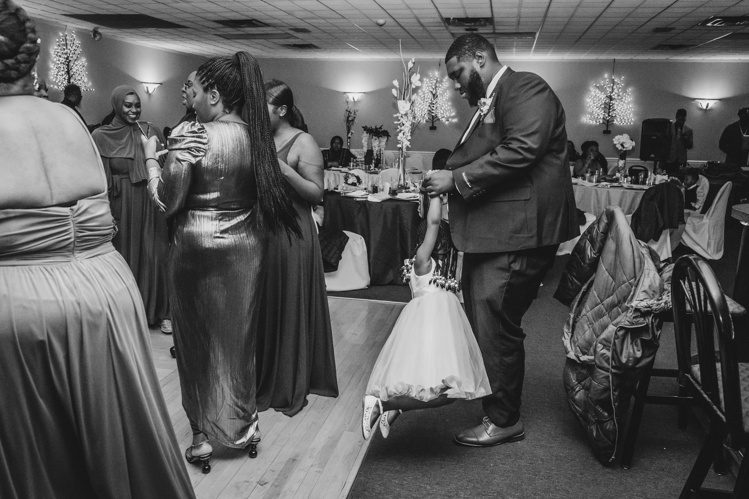 A man in a suit holding a young girl's hand during a formal event, with women dressed in gowns dancing and socializing in the background at a decorated banquet hall.