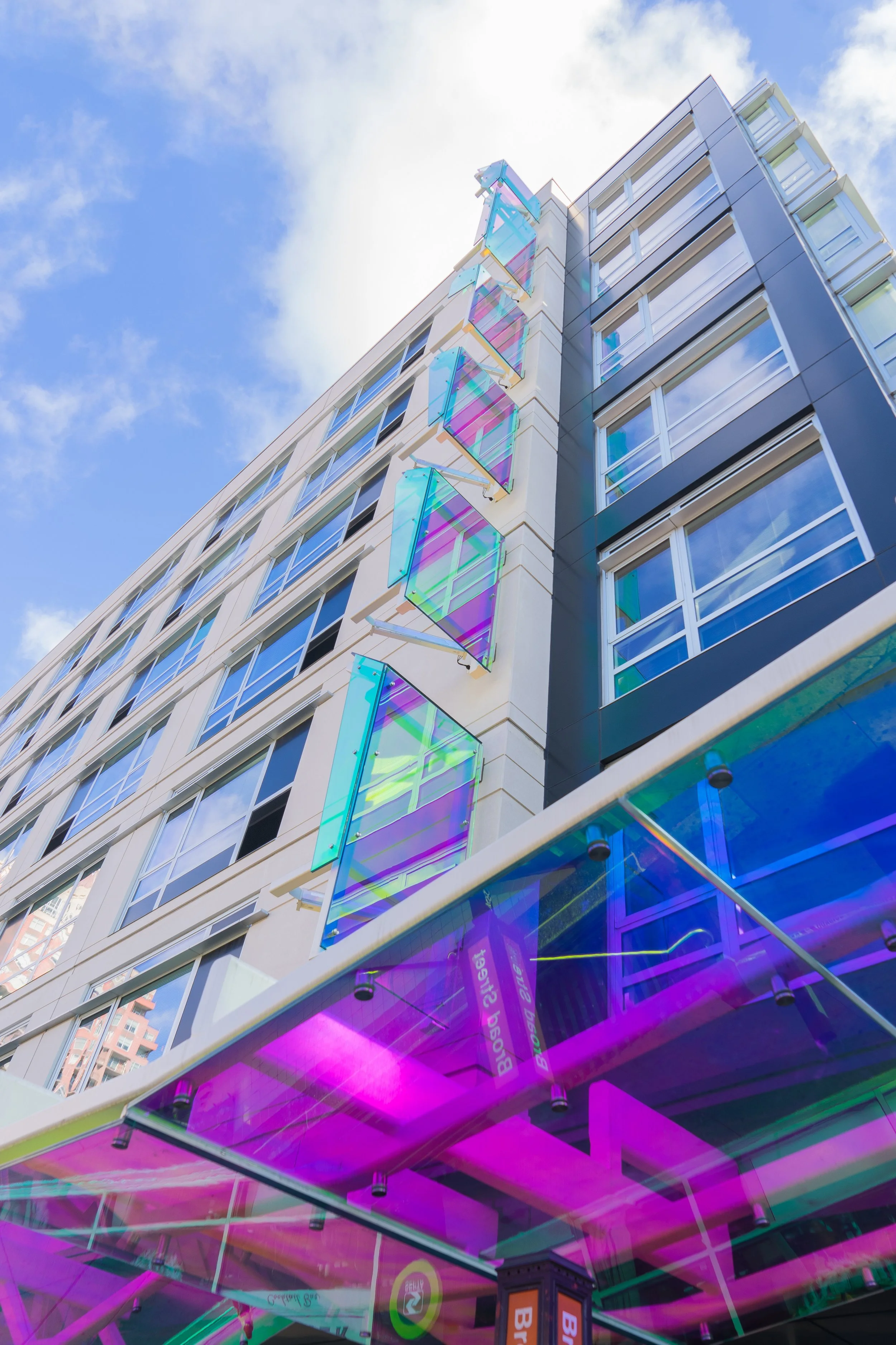 A modern multi-story building with colorful, iridescent window panels and a pink and purple neon-lit storefront at street level.