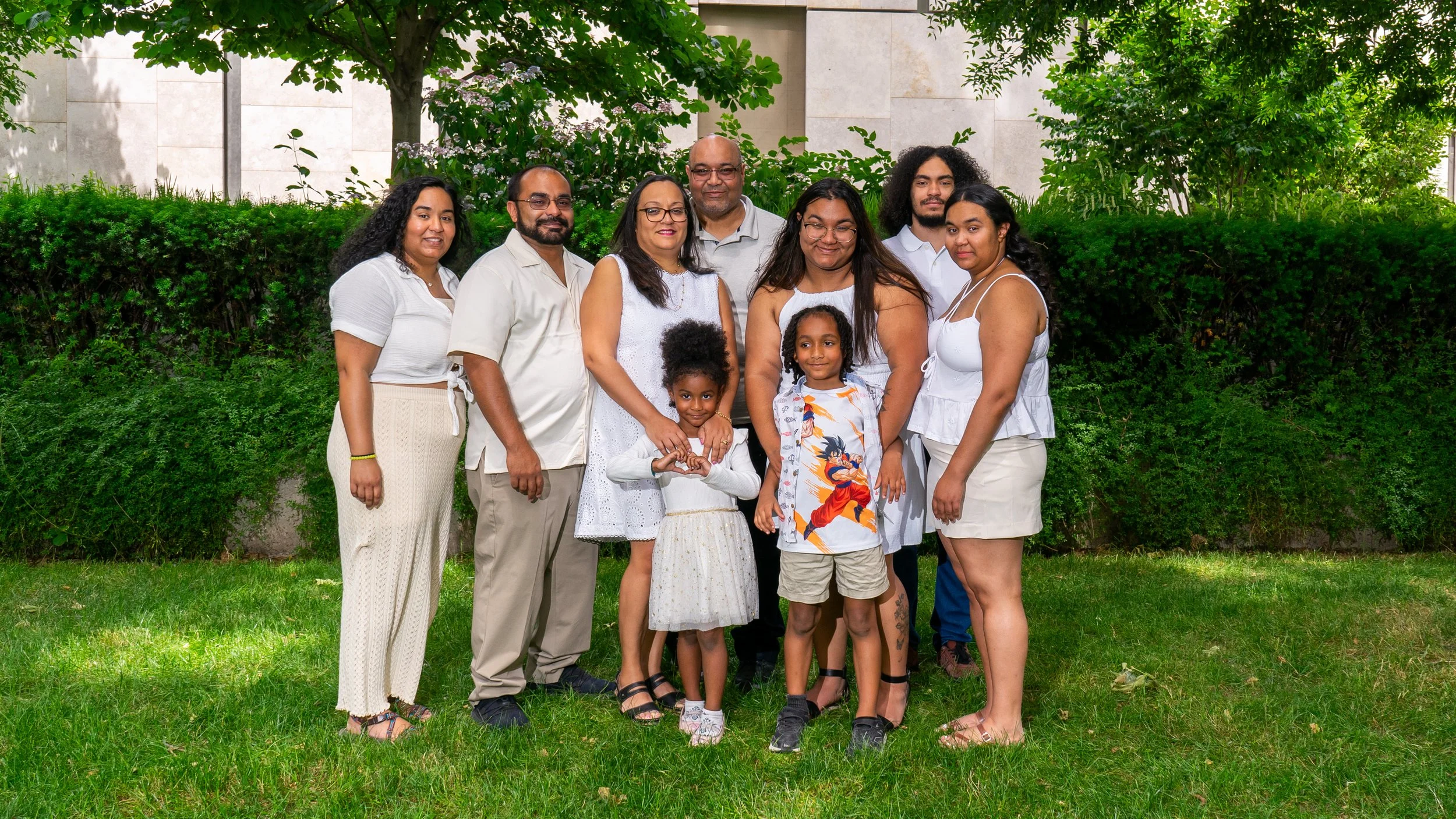 A group of ten people, including children, posing outdoors on a lawn with green bushes and trees in the background. All are dressed in light-colored casual summer clothing, smiling and standing close together.