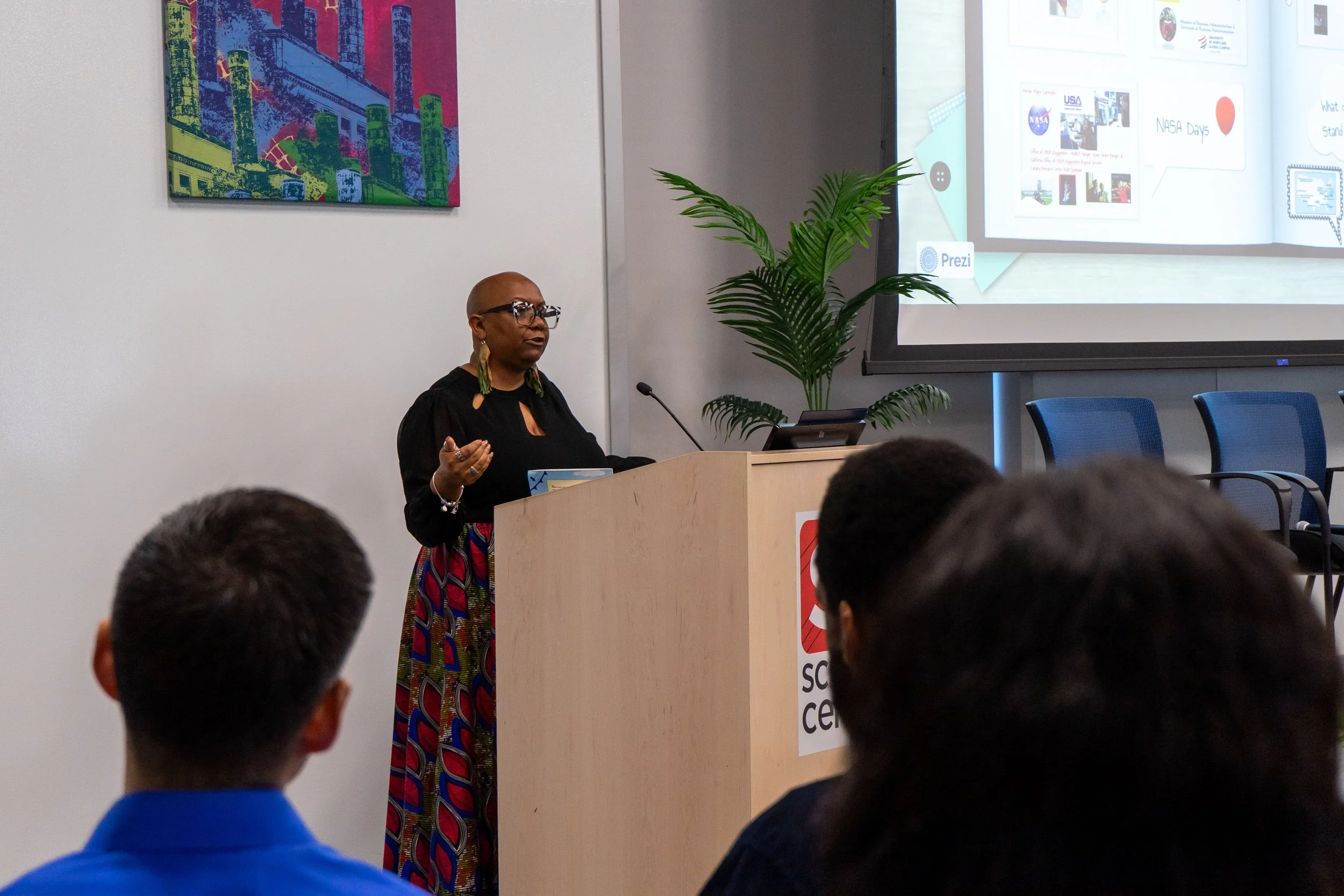 A woman with glasses and colorful earrings giving a presentation at a podium in front of an audience. A screen to her right displays a presentation slide, and a large potted plant is beside her.