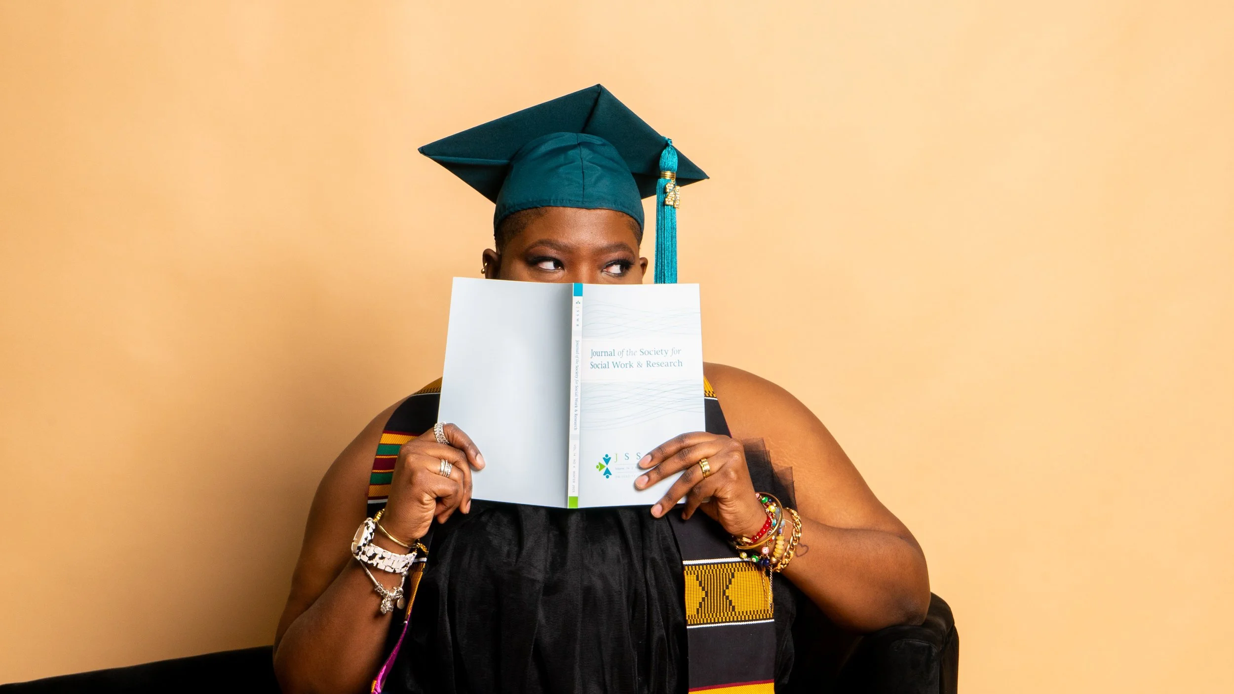 A woman in graduation attire holding a book in front of her face, looking to the side, against a beige background.