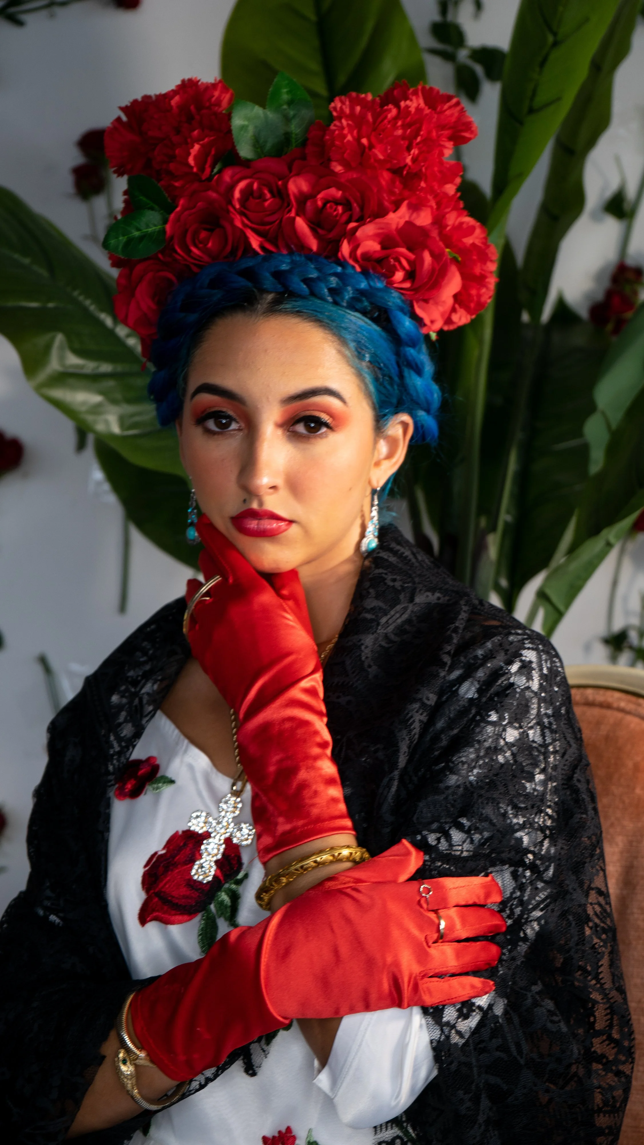 A woman with blue braided hair, wearing red gloves, a black lace shawl, and jewelry, poses in front of large green leaves and red roses.