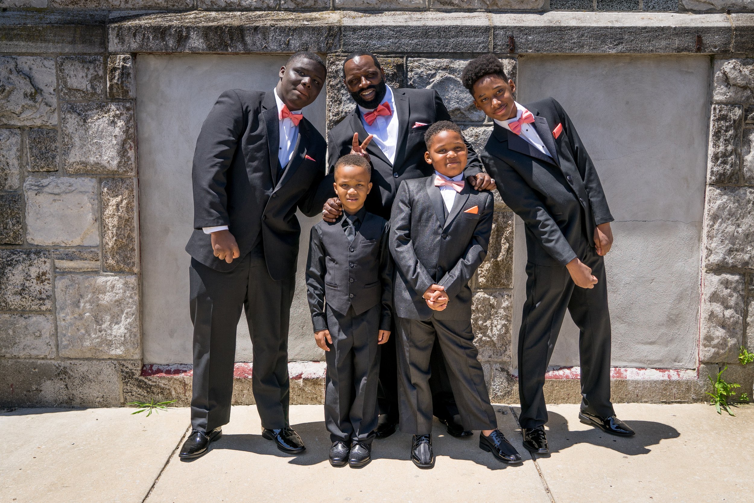 A group of six people, including one adult and five boys, dressed in formal attire with black suits and pink bow ties, posing outdoors in front of a stone wall.