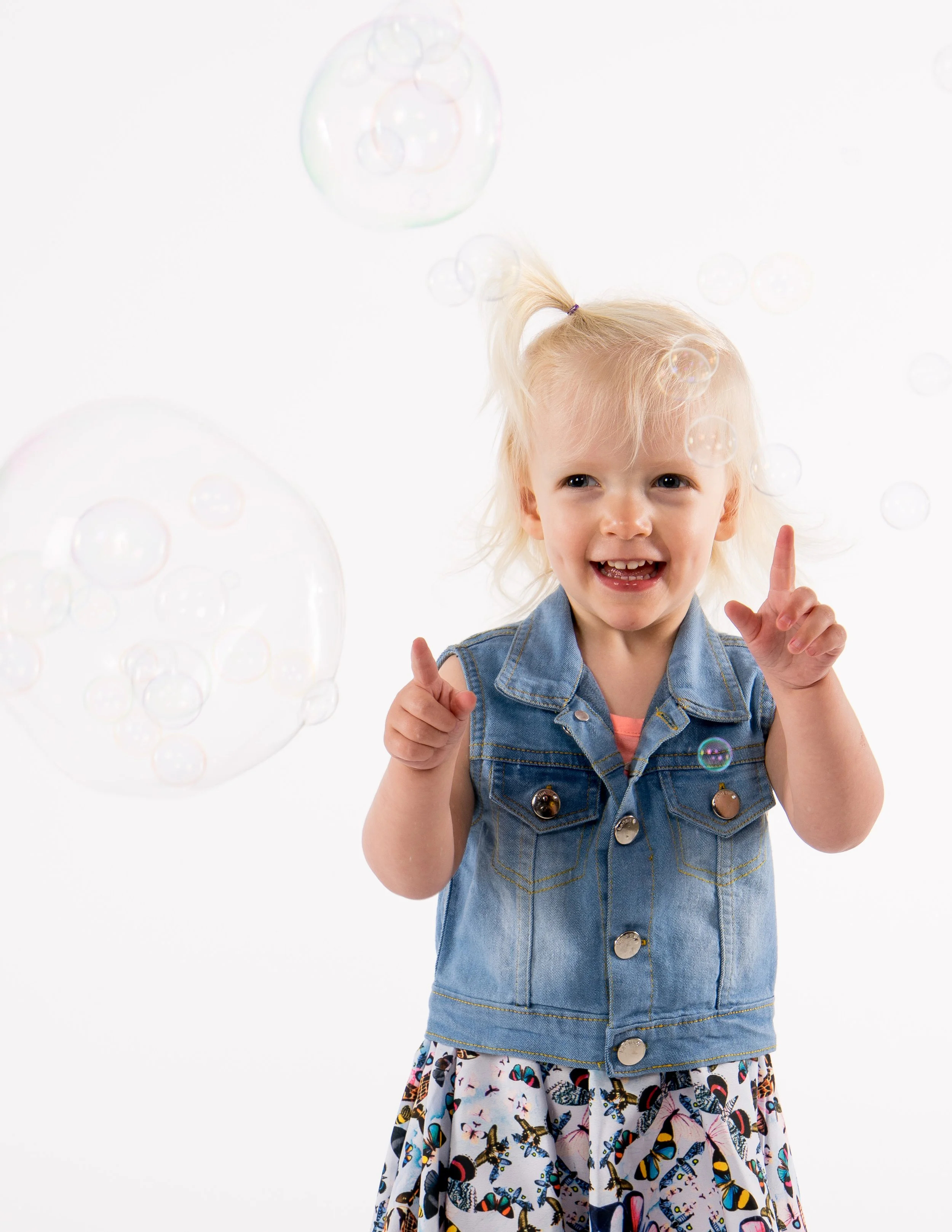 A young girl with blonde hair in a ponytail, wearing a denim vest and a butterfly-patterned skirt, is playing with soap bubbles against a white background.