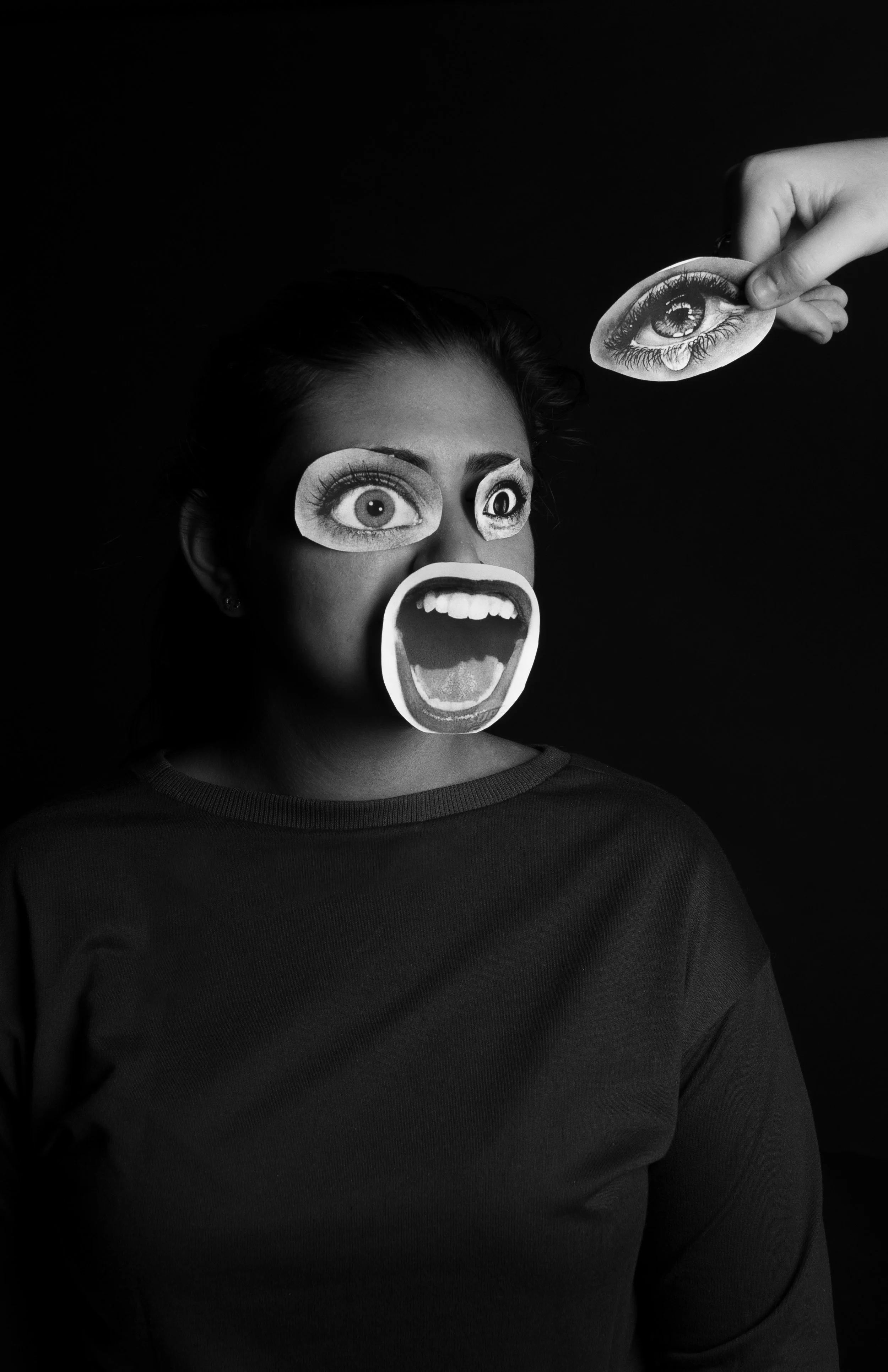 Black and white photo of a woman with cutouts of eyes and a mouth on her face, and a hand holding a cutout of an eye above her head.