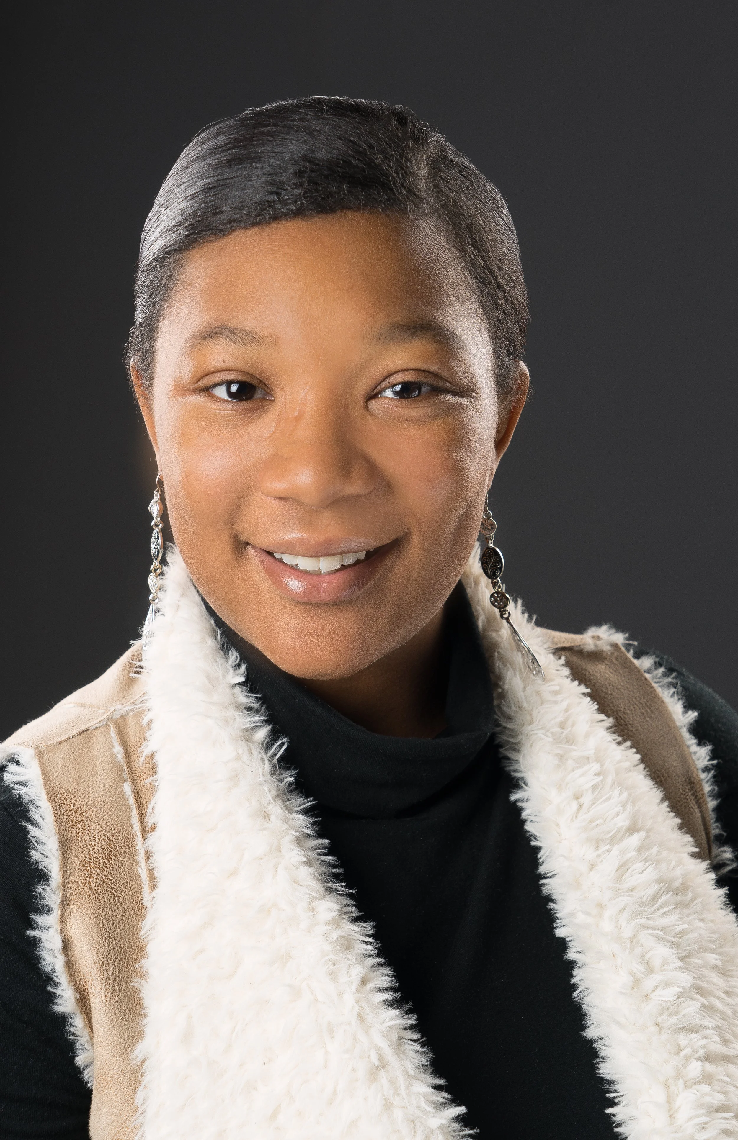 Headshot of a woman with short, dark hair, smiling, wearing earrings, a black turtleneck, and a beige jacket with fur lining.