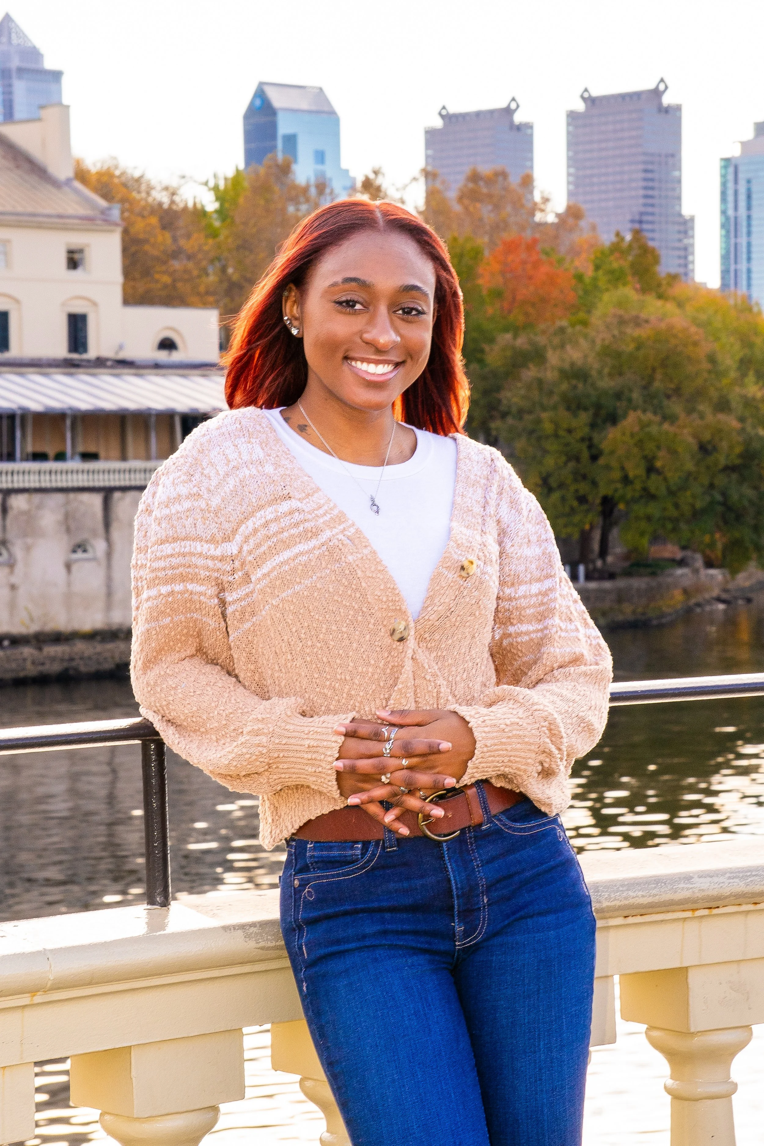 A young woman with red hair smiling outdoors near a river, wearing a beige cardigan, white shirt, and blue jeans with autumn trees and city buildings in the background.