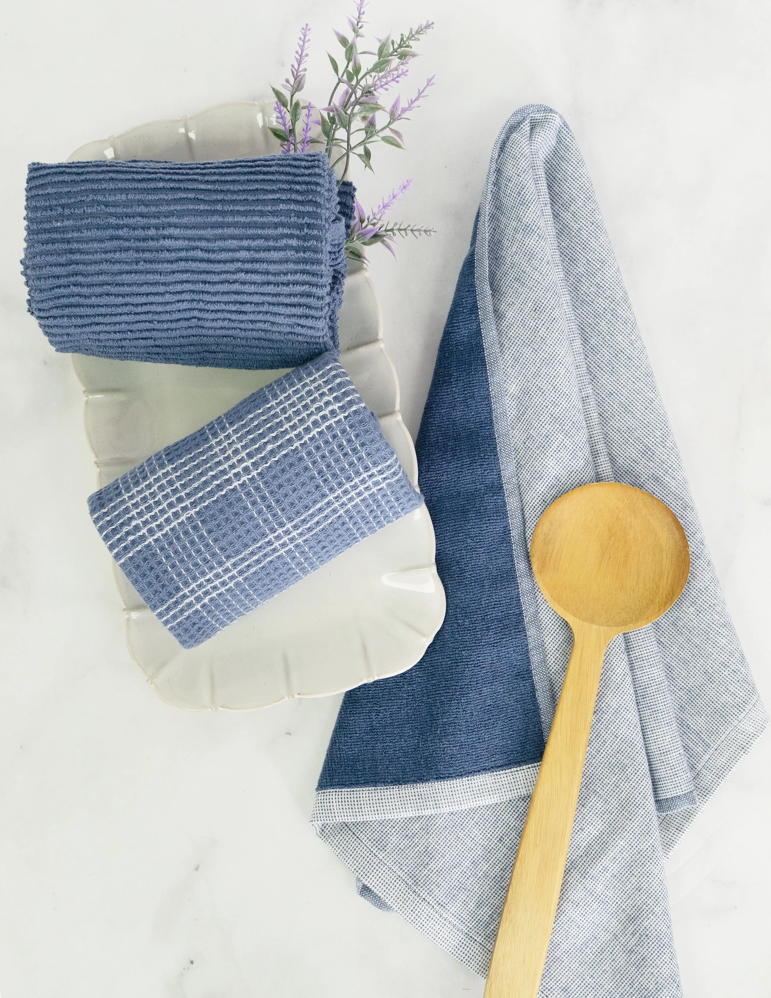 Blue and gray kitchen towels on a white ceramic dish, wooden spoon, and purple flowers on a white surface.