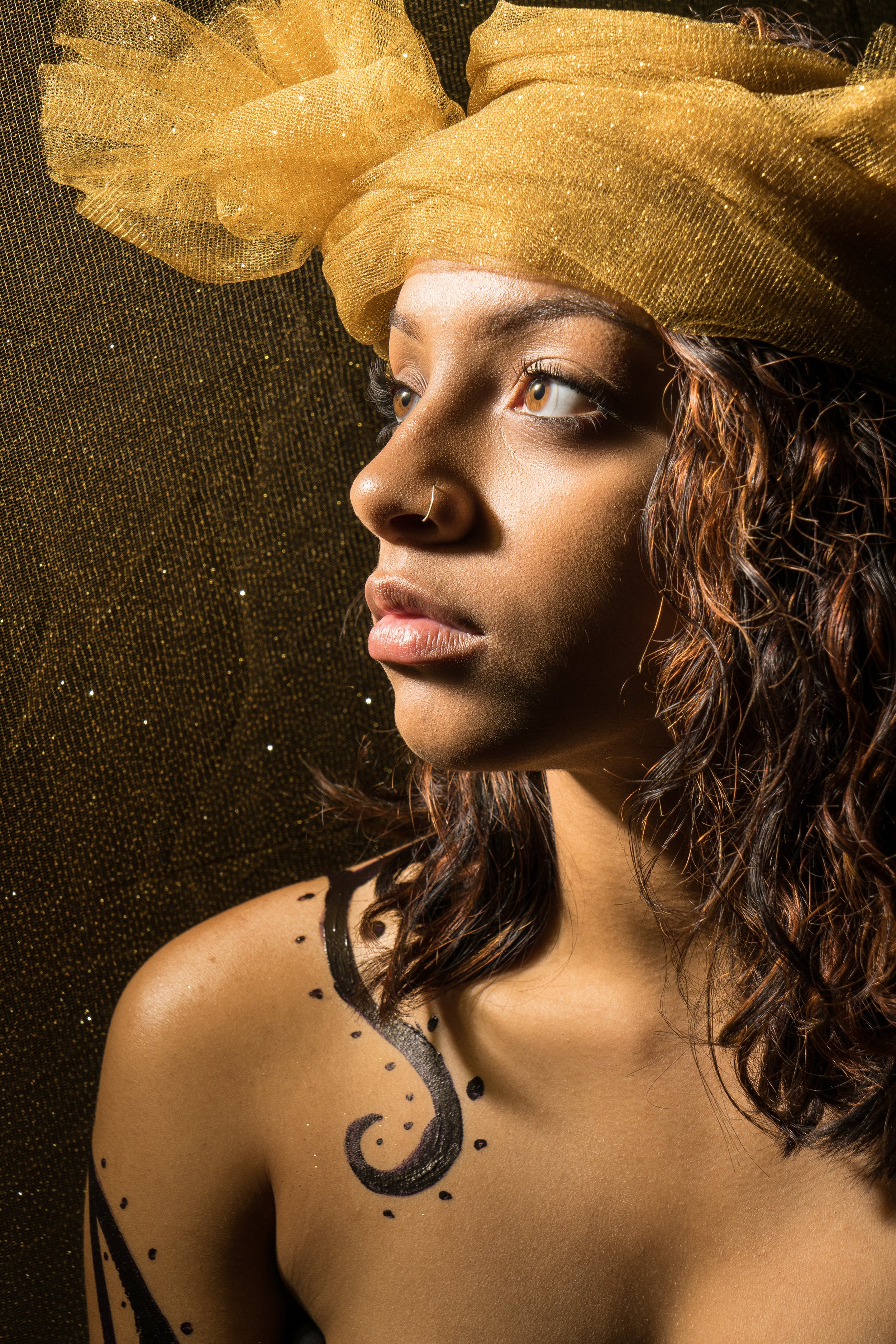A young woman with curly brown hair wears a large gold headwrap and has black decorative tattoos on her shoulder and collarbone. She looks to the right with a serious expression, against a glittery gold background.