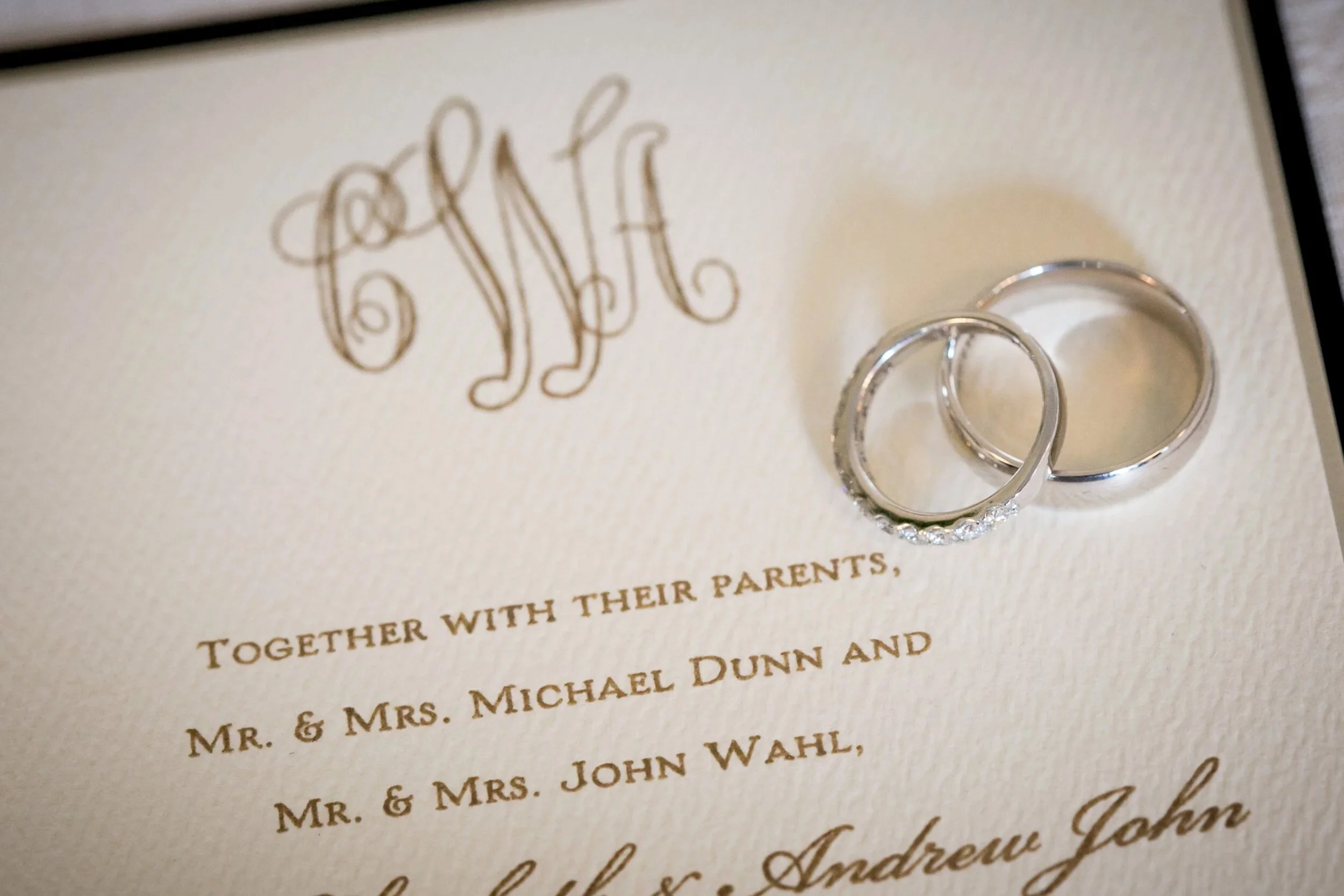 Close-up of a wedding invitation with two wedding rings placed on it. The invitation has a monogram at the top and text in gold and black describing the event.