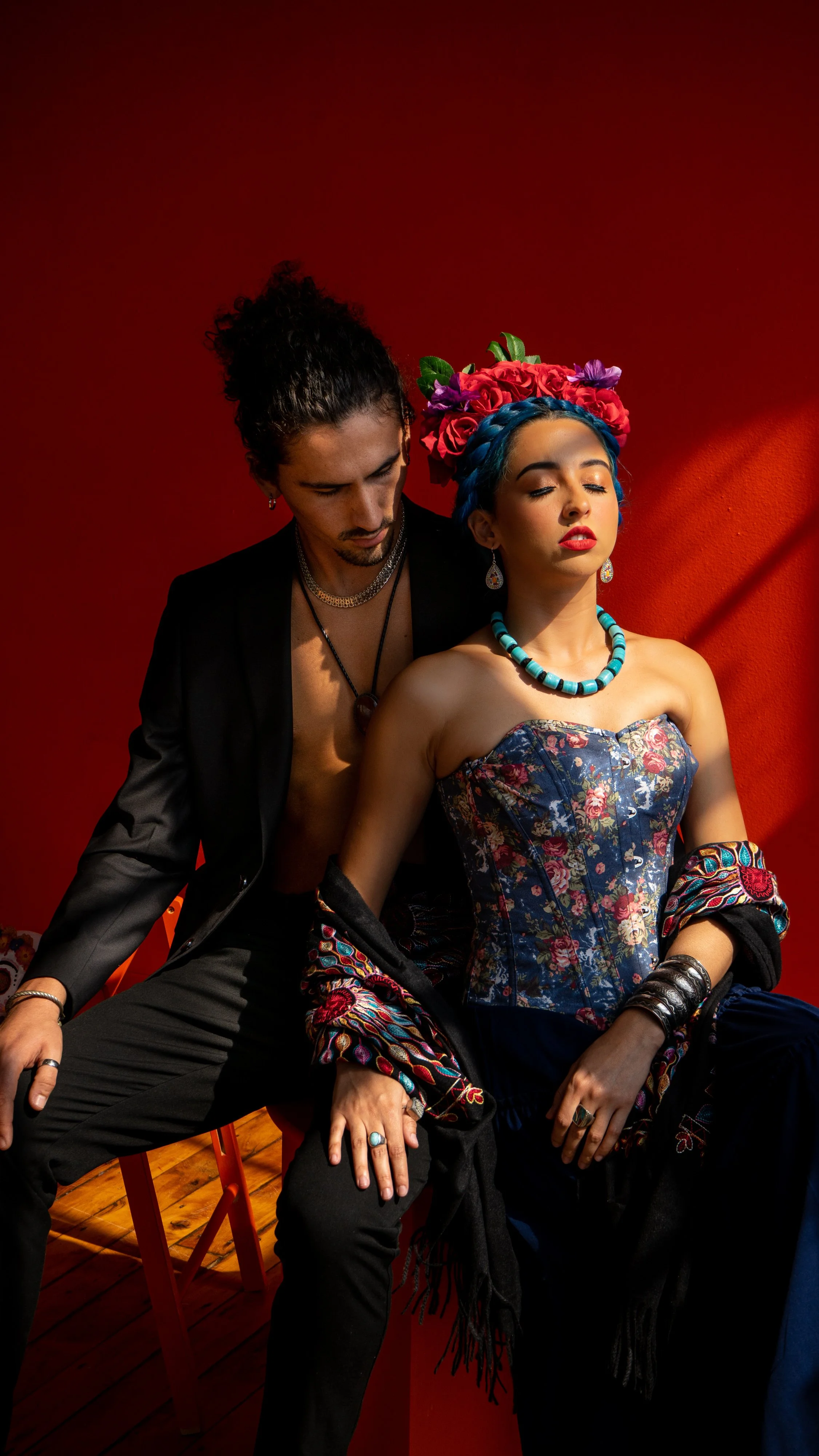 A man and a woman seated against a red wall. The woman wears a floral corset, vibrant jewelry, and a flower crown, while the man wears a black jacket, necklace, and dark pants, both with closed eyes and contemplative expressions.