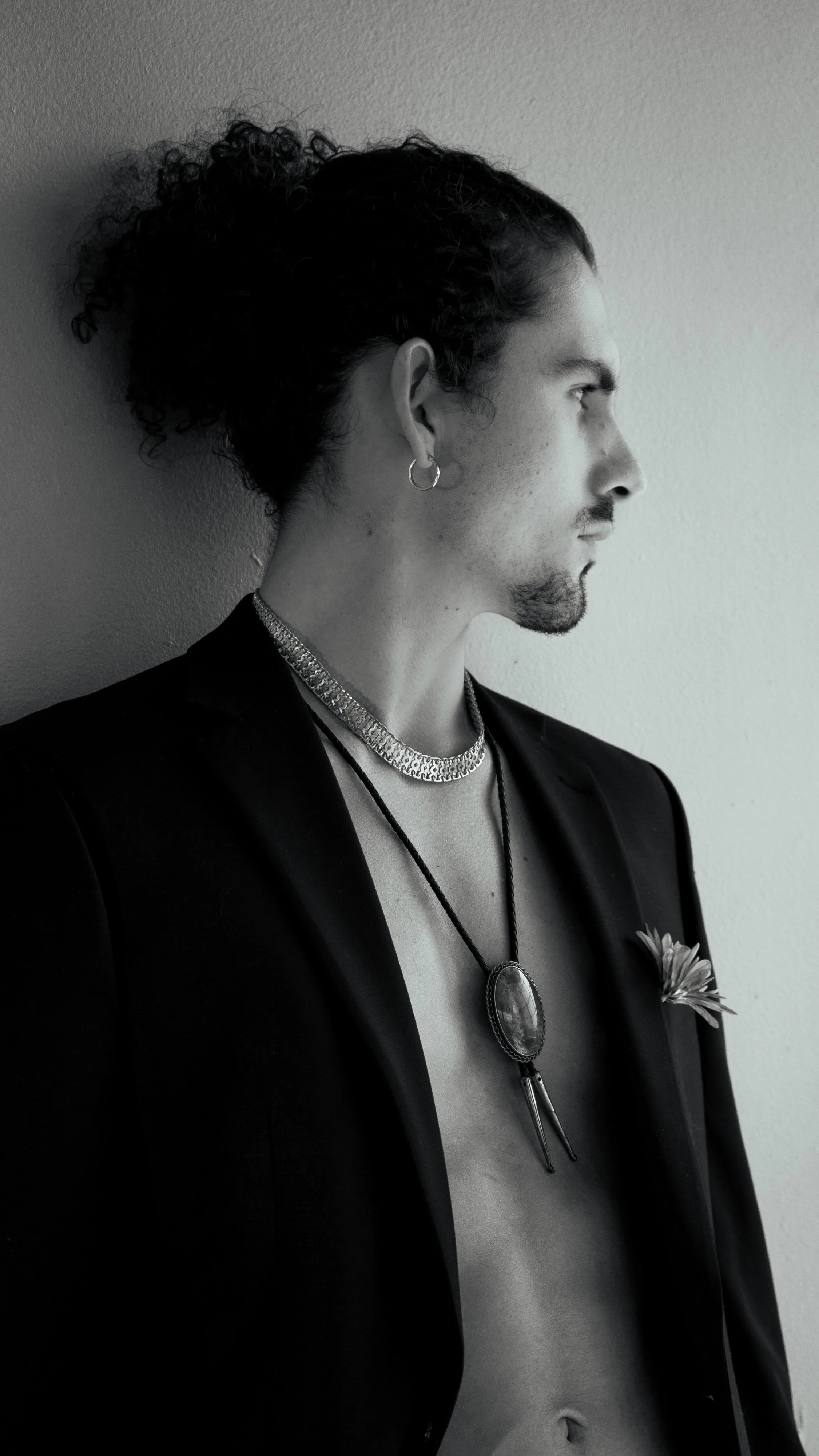 A person with curly hair leaning against a wall, wearing jewelry including a necklace, earrings, and a brooch, with a blazer open to reveal their bare chest and abdomen, in black and white.